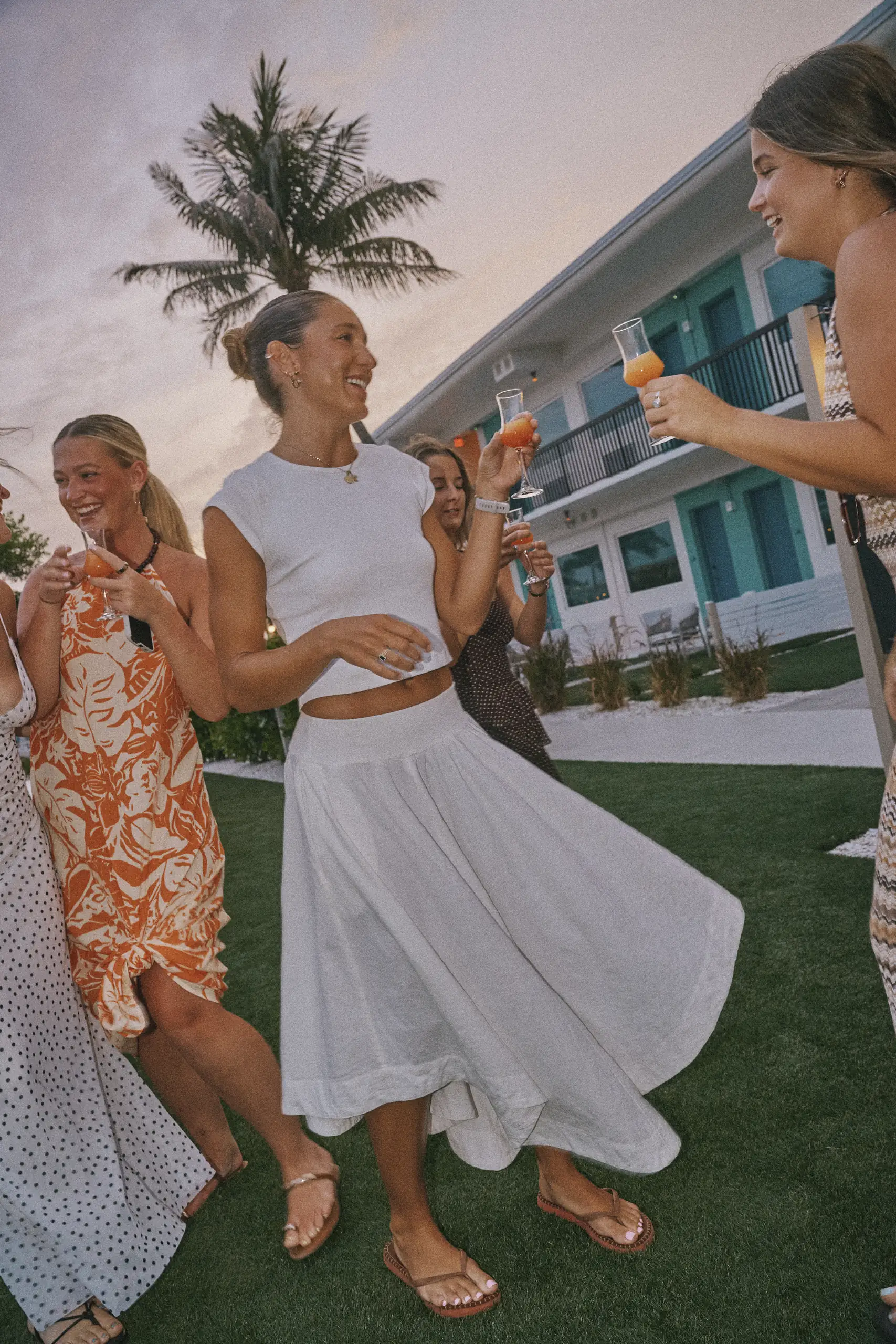 Group of women dancing outdoors at sunset, holding drinks and smiling near a palm tree and building.