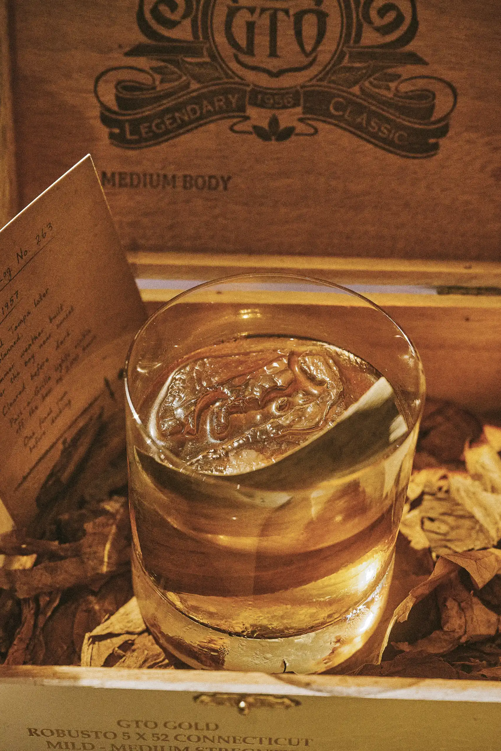 Glass with a large ice cube and a drink inside a wooden cigar box with dried tobacco leaves.
