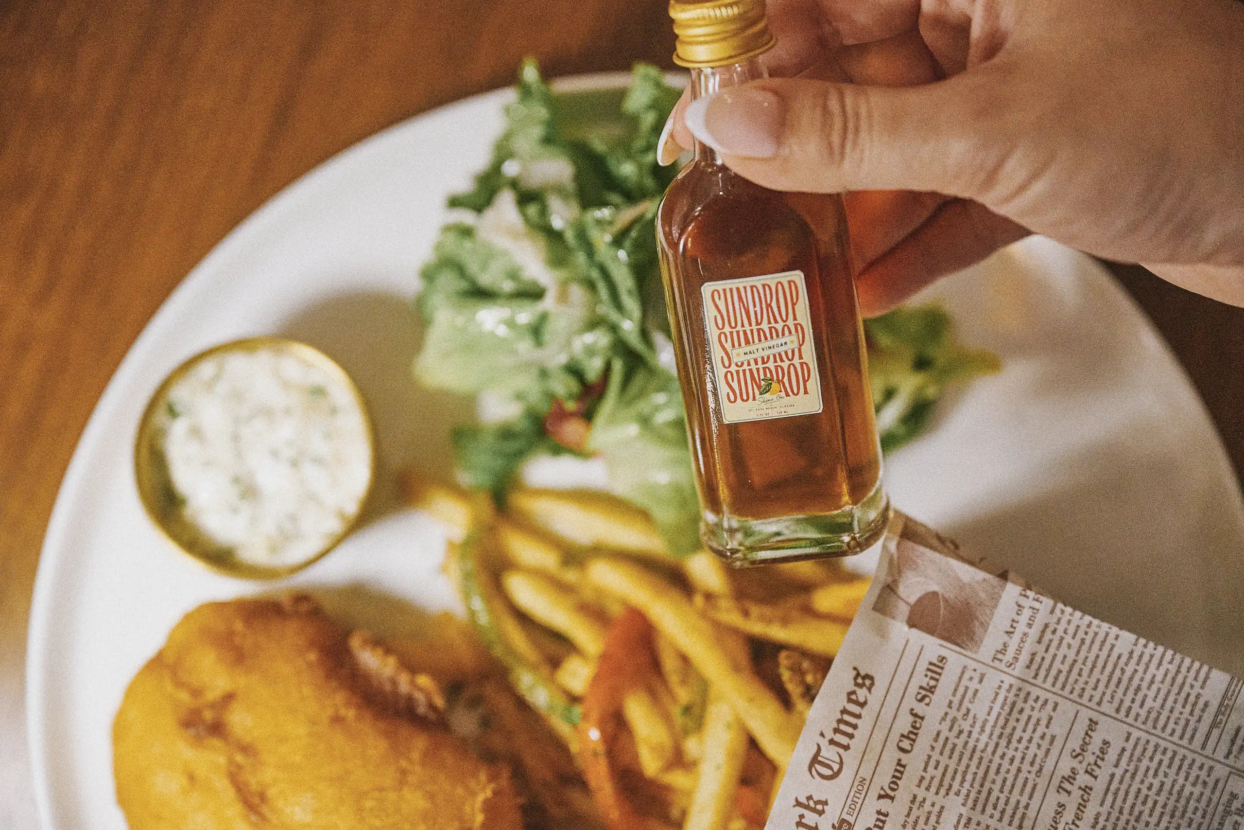 Hand holding small bottle of Sundrop malt vinegar over fish, fries, salad, and sauce on a white plate.