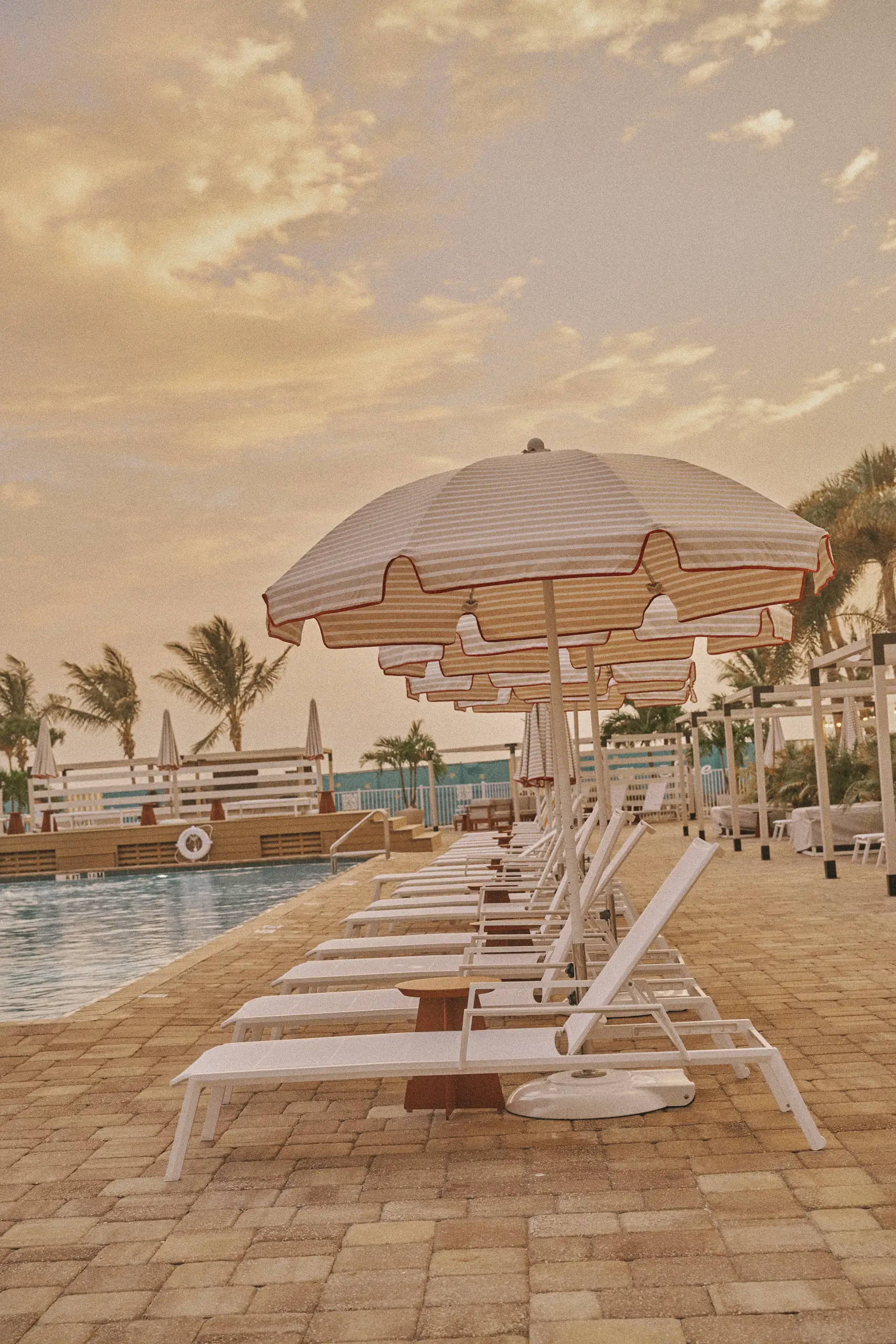 A row of white poolside lounge chairs under striped umbrellas with palm trees and cloudy sky.