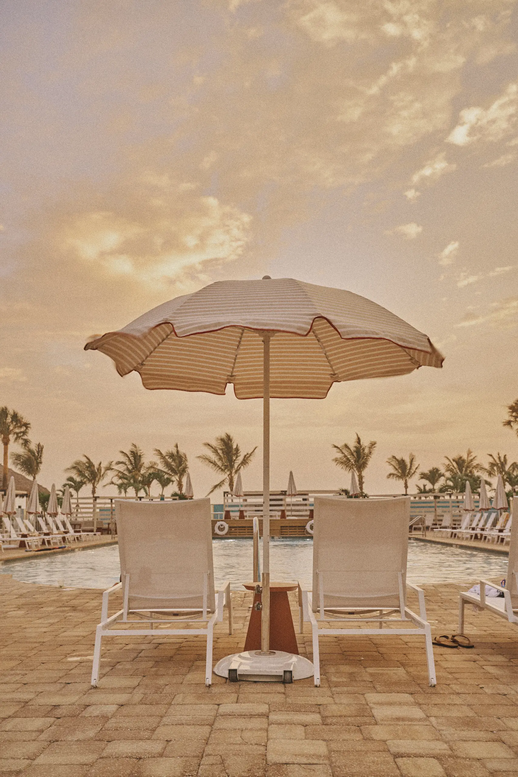 Two white lounge chairs under a striped umbrella by a pool with palm trees at sunset.
