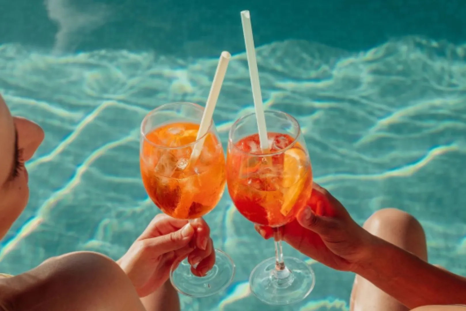 Two people clinking glasses of Aperol Spritz with straws at the edge of a sparkling pool.