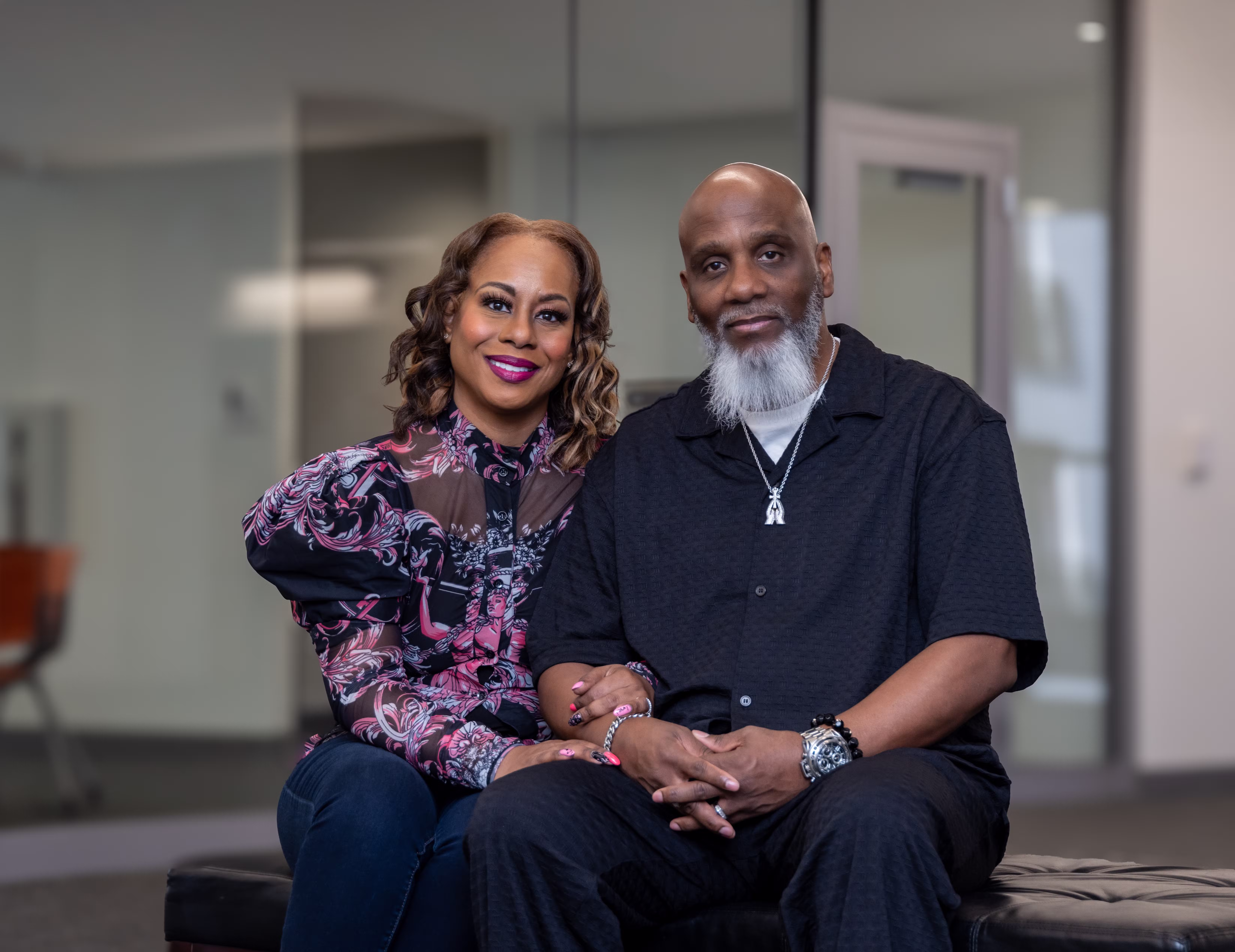 Smiling couple seated together indoors, the woman wearing a patterned puffed sleeve blouse and the man in a black shirt with a white beard.