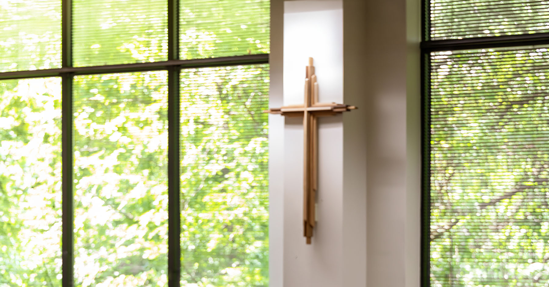 Indoor chapel scene with a wooden cross mounted on the wall and a table covered with a white cloth in front of large windows with green foliage outside.
