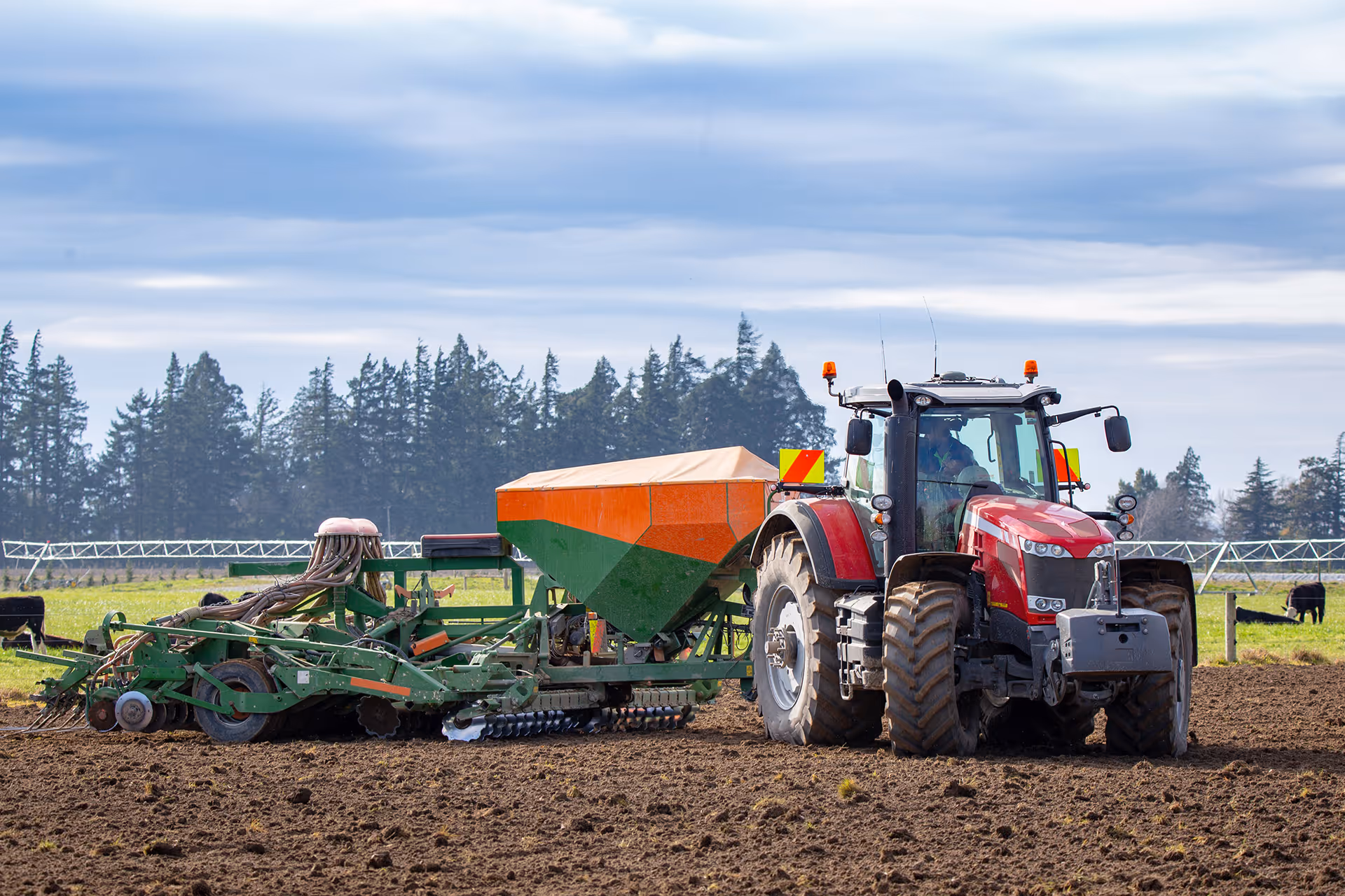 Red tractor with green and orange seed drill attachment working on a plowed farm field.