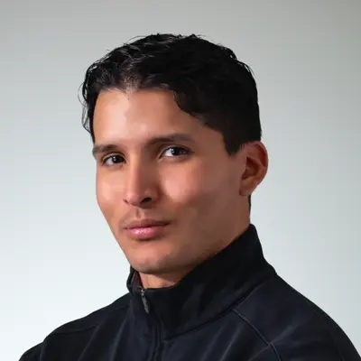 Portrait of a man with short dark hair wearing a black zip-up jacket against a gray background.
