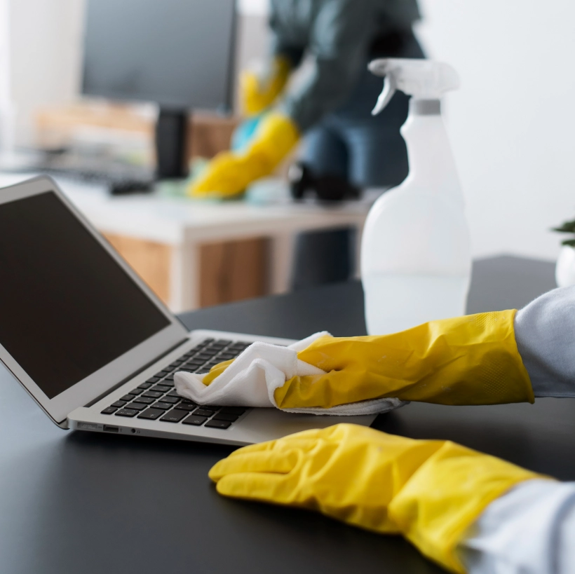 Cleaner in yellow gloves disinfecting a laptop and desk during office cleaning