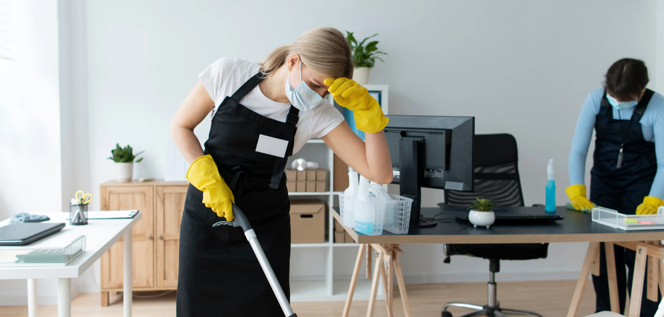 Two cleaners in aprons and gloves cleaning desks and floors in a bright suburban office