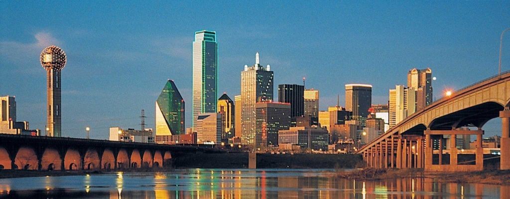 Dallas skyline at sunset with a river and two bridges reflecting city lights.