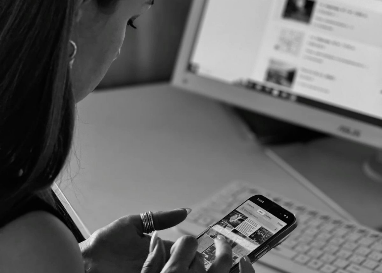 Woman using a smartphone while sitting at a desk with a computer keyboard and monitor in the background.