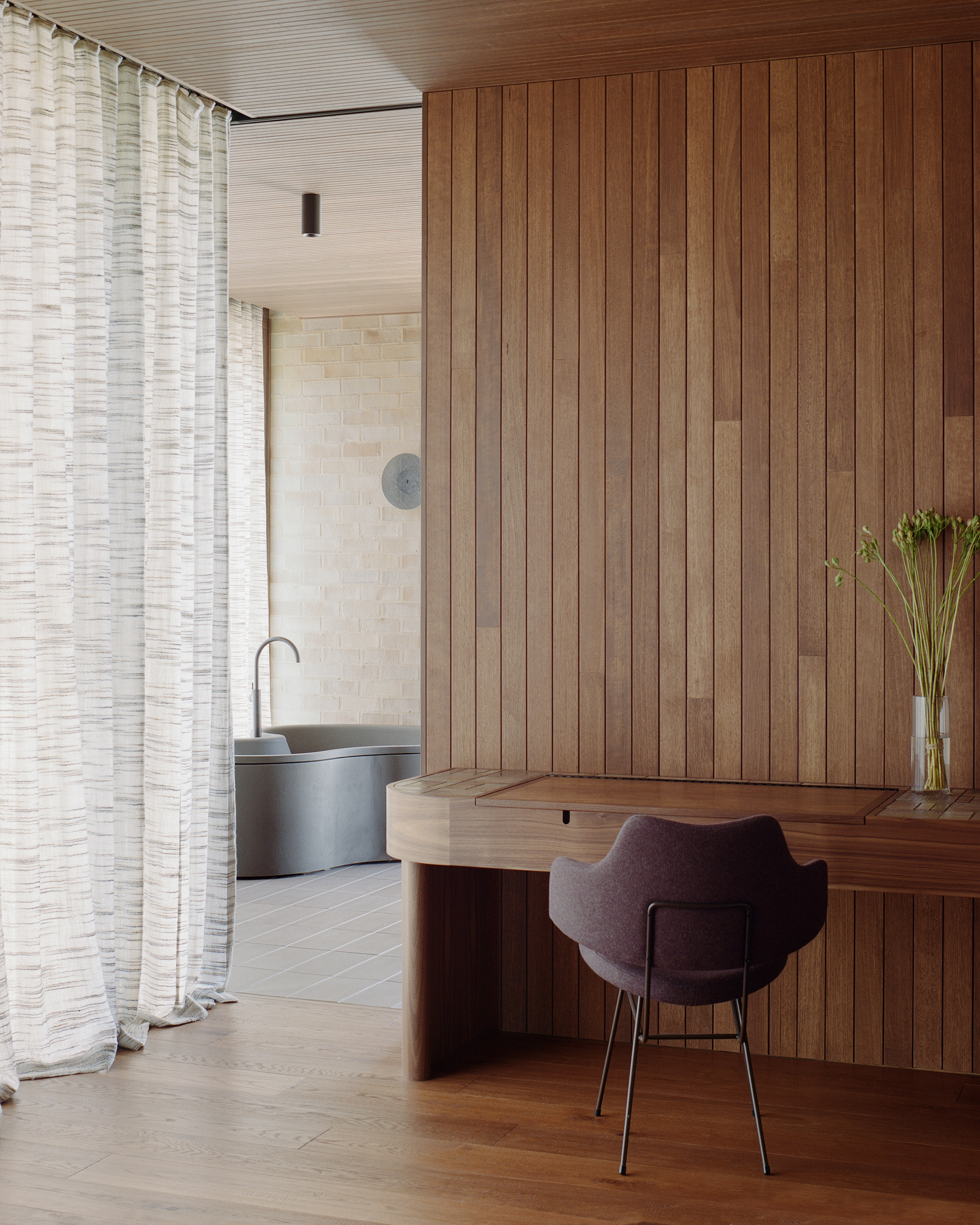 Luxury main bedroom suite featuring a built-in timber vanity desk and a modern purple armchair against a wood-paneled wall. In the background, a curved grey freestanding bathtub is visible behind sheer, textured floor-to-ceiling curtains.