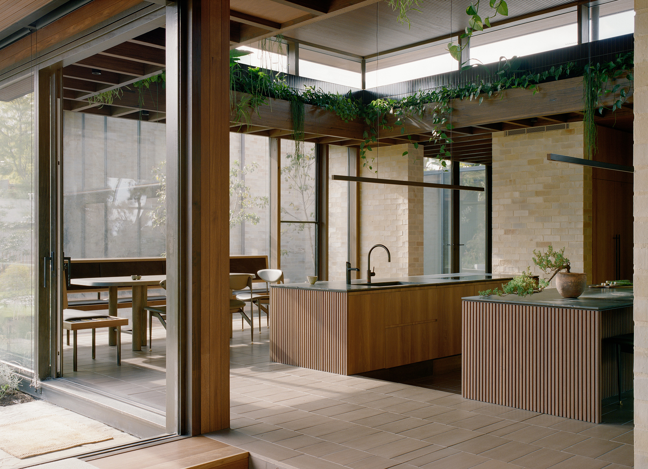 Open-plan modern kitchen and dining room opening onto an outdoor area through large sliding glass doors. Features twin ribbed timber island benches, integrated indoor plant troughs, and a built-in dining banquet booth in the background.