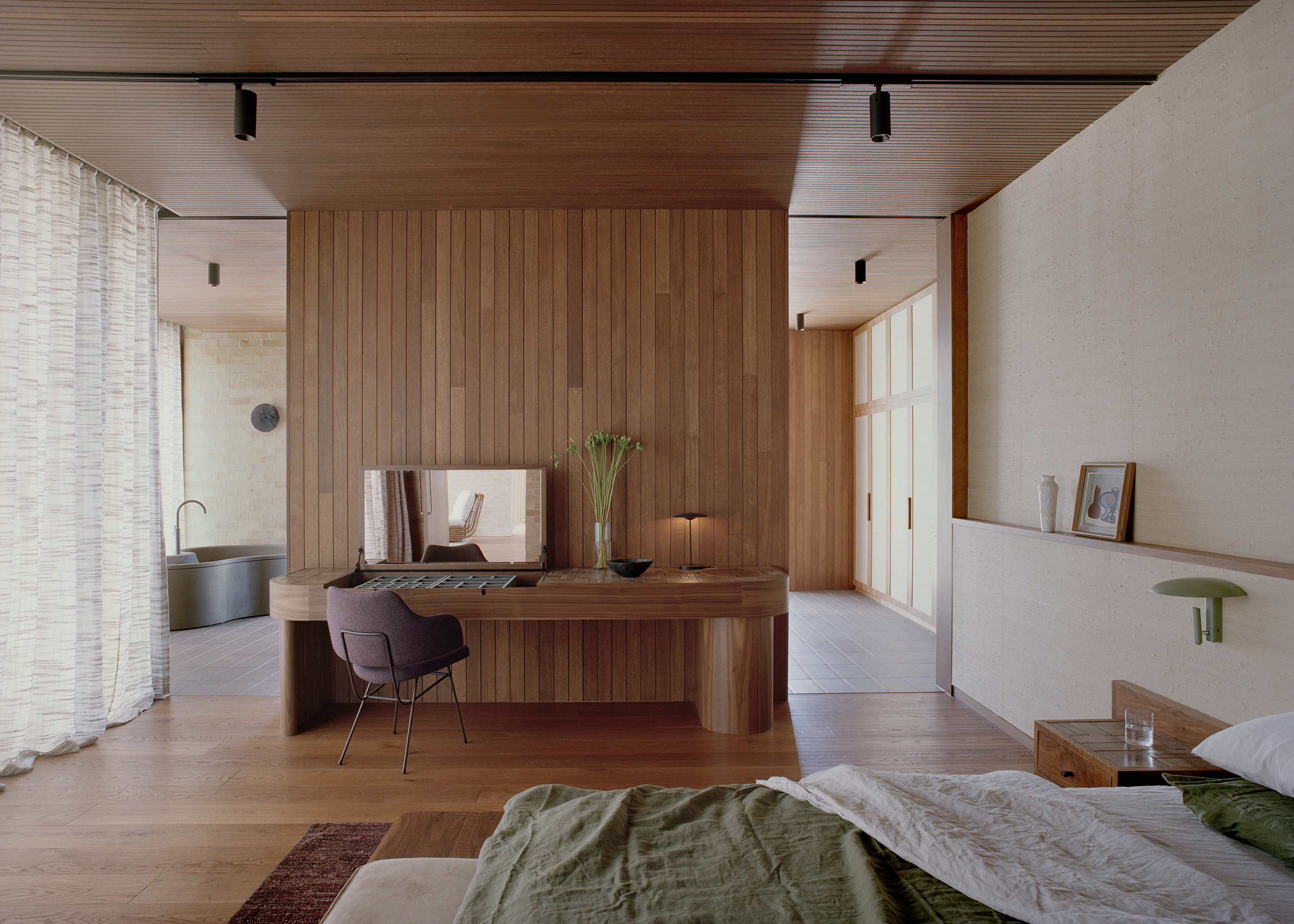 Spacious master bedroom interior showing a low-profile bed with olive green linen. A large central timber wall partition separates the sleeping area from a dressing vanity with an integrated mirror and an open ensuite bathroom beyond.