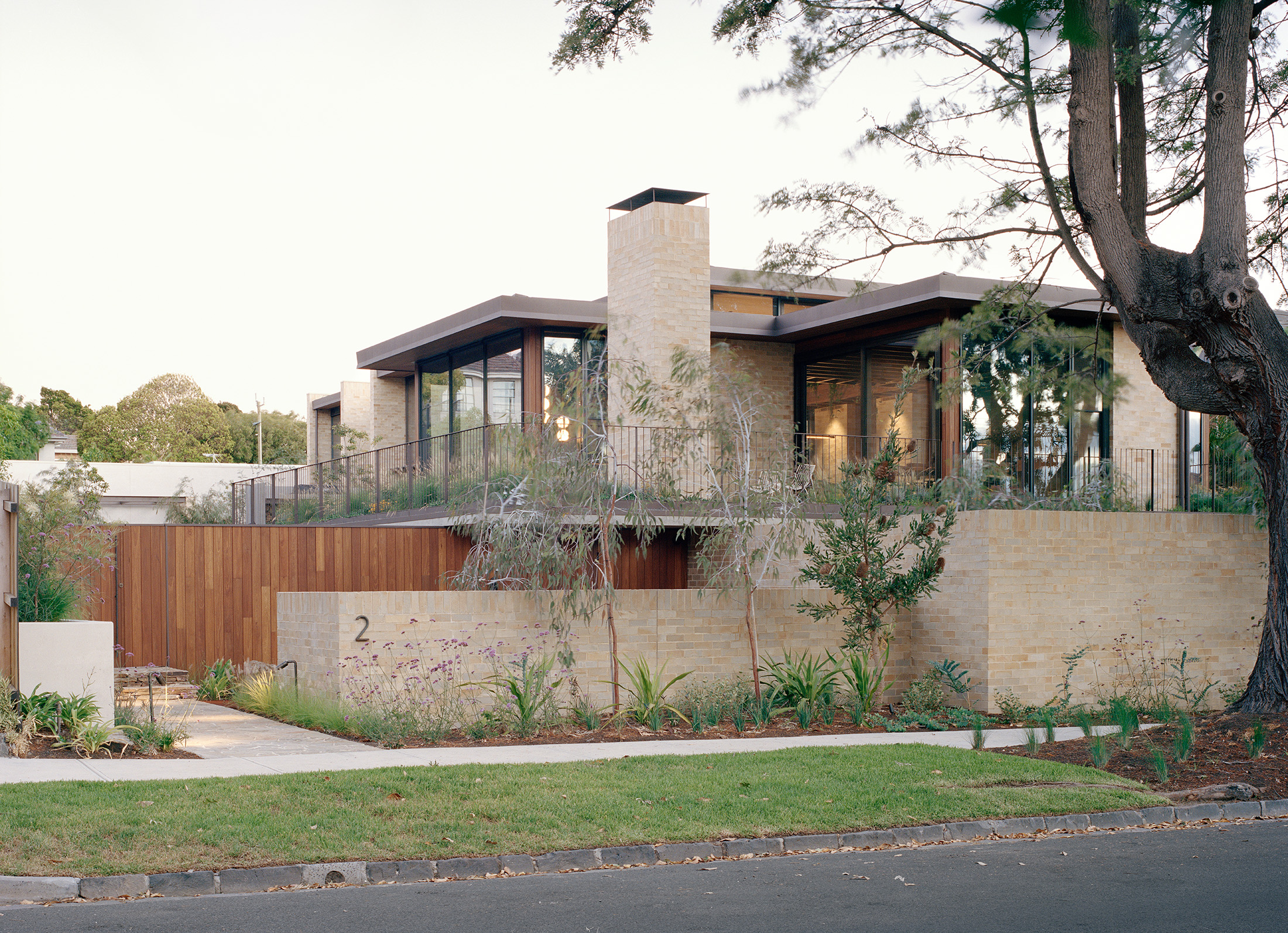 Street view of a contemporary luxury home designed by Archier. The architecture combines light brick walls, timber paneling, and an upper-level glass pavilion. A minimalist front garden with native plants lines the concrete path and driveway.
