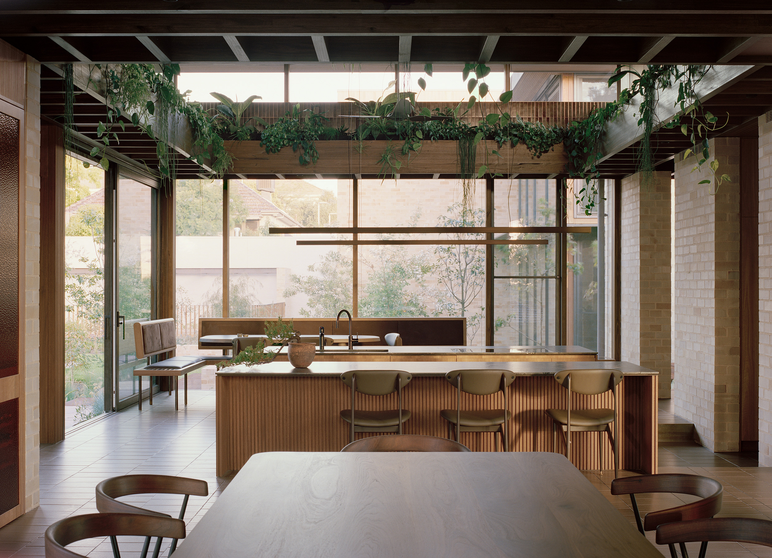 Mid-century modern kitchen interior by Archier featuring a ribbed timber island bench, minimalist bar stools, and polished tile flooring. An integrated overhead wooden planter box with trailing indoor plants runs beneath a high clerestory window.
