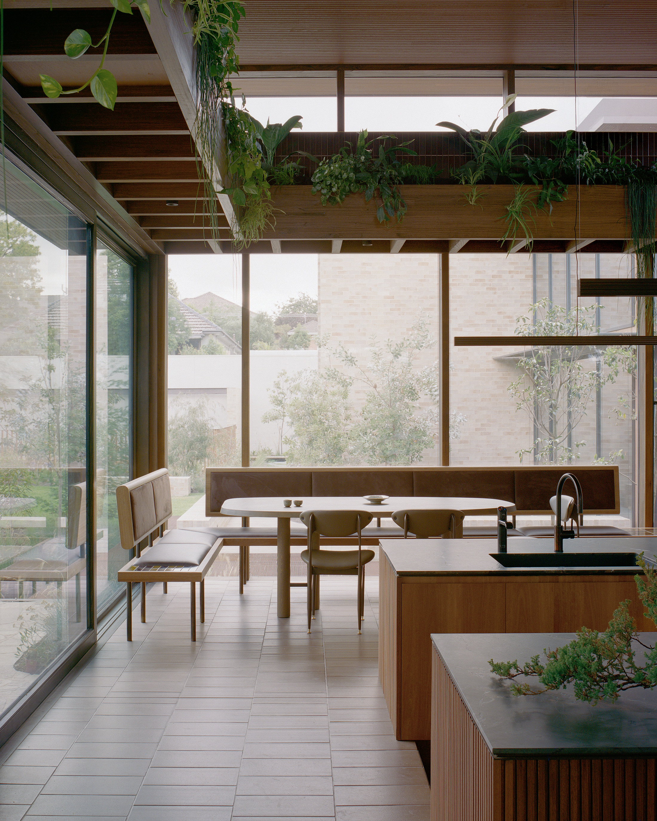 Minimalist dining area featuring a custom built-in leather banquet booth and an oval timber table. Floor-to-ceiling glass windows look out to a courtyard garden, while overhead wooden beams house trailing green indoor plants.