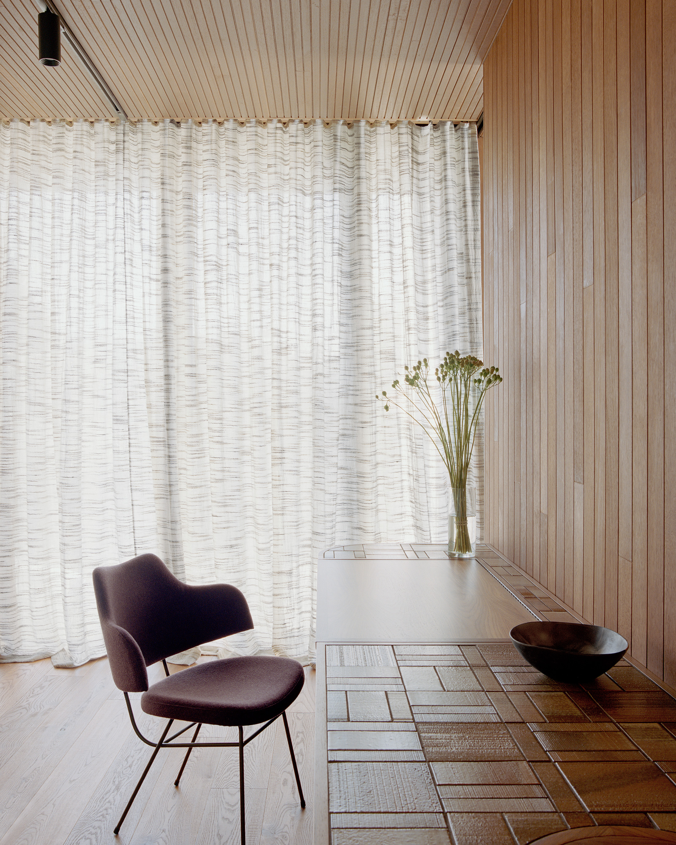 Detail view of a custom mid-century style timber desk featuring a unique tiled wood surface pattern. A minimalist dark purple desk chair sits beside the workspace, framed by soft, sheer textured curtains filtering natural light.