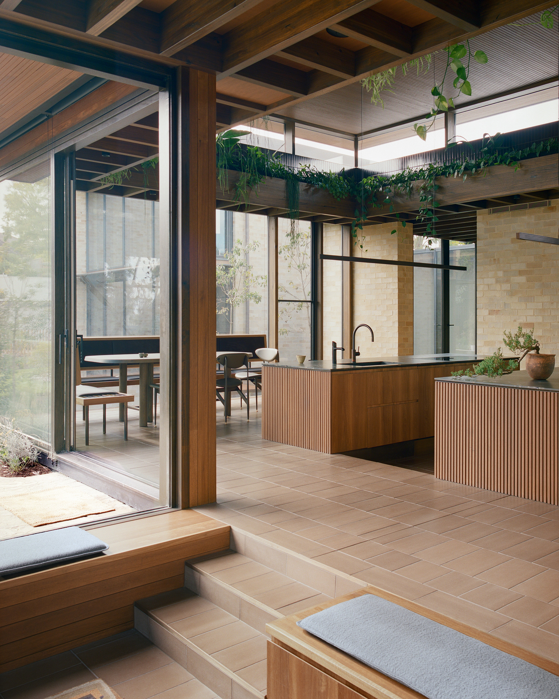 Contemporary interior design highlighting a split-level transition with low timber bench seating and grey cushions. The space opens up to a kitchen with ribbed wood cabinetry, tile floors, exposed timber ceiling beams, and indoor greenery.