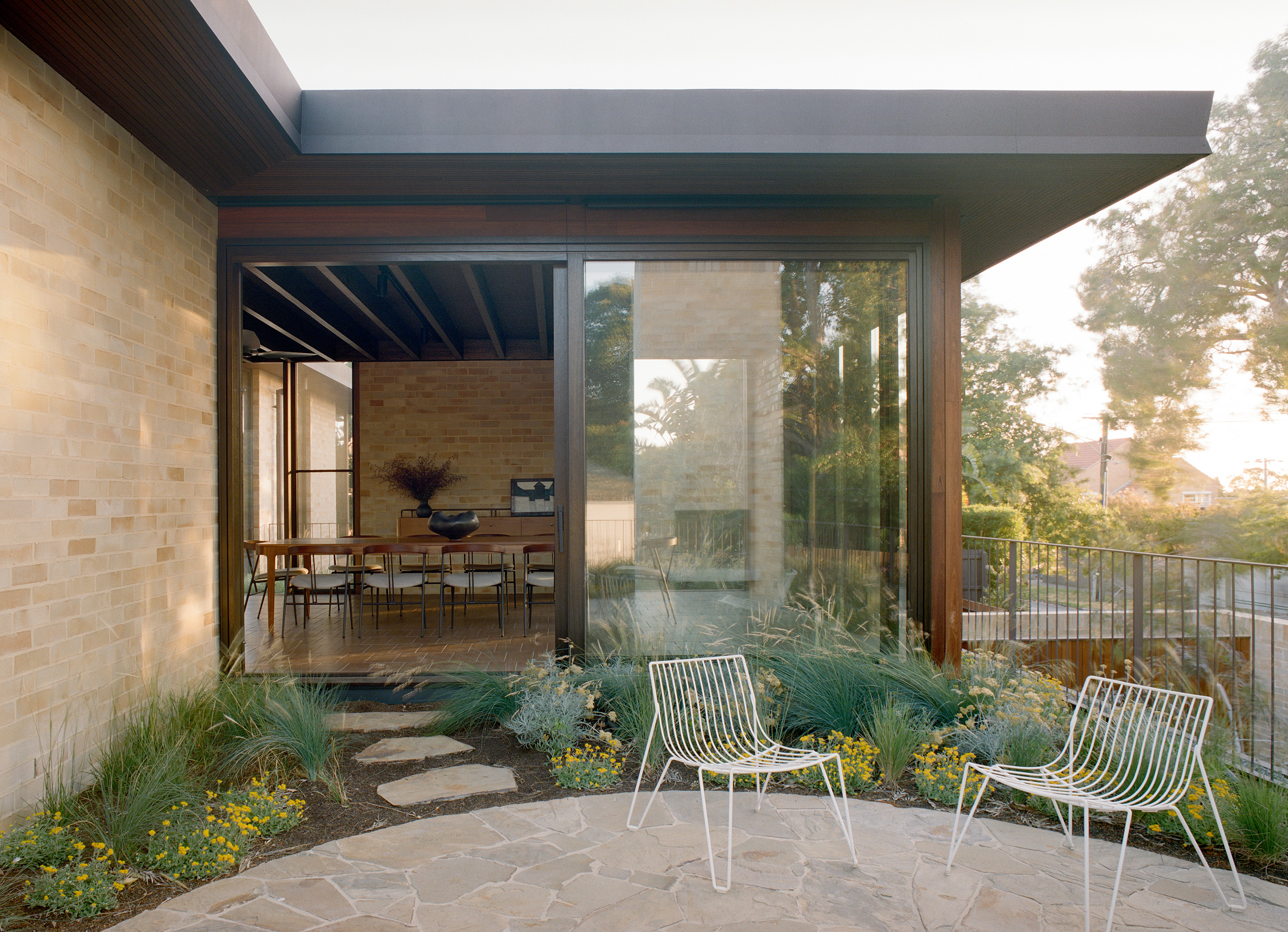 Indoor-outdoor living space featuring sliding glass doors opened to an open-plan dining room. Outside, a flagstone patio features two white wire-frame outdoor chairs surrounded by native grasses and yellow flowers.