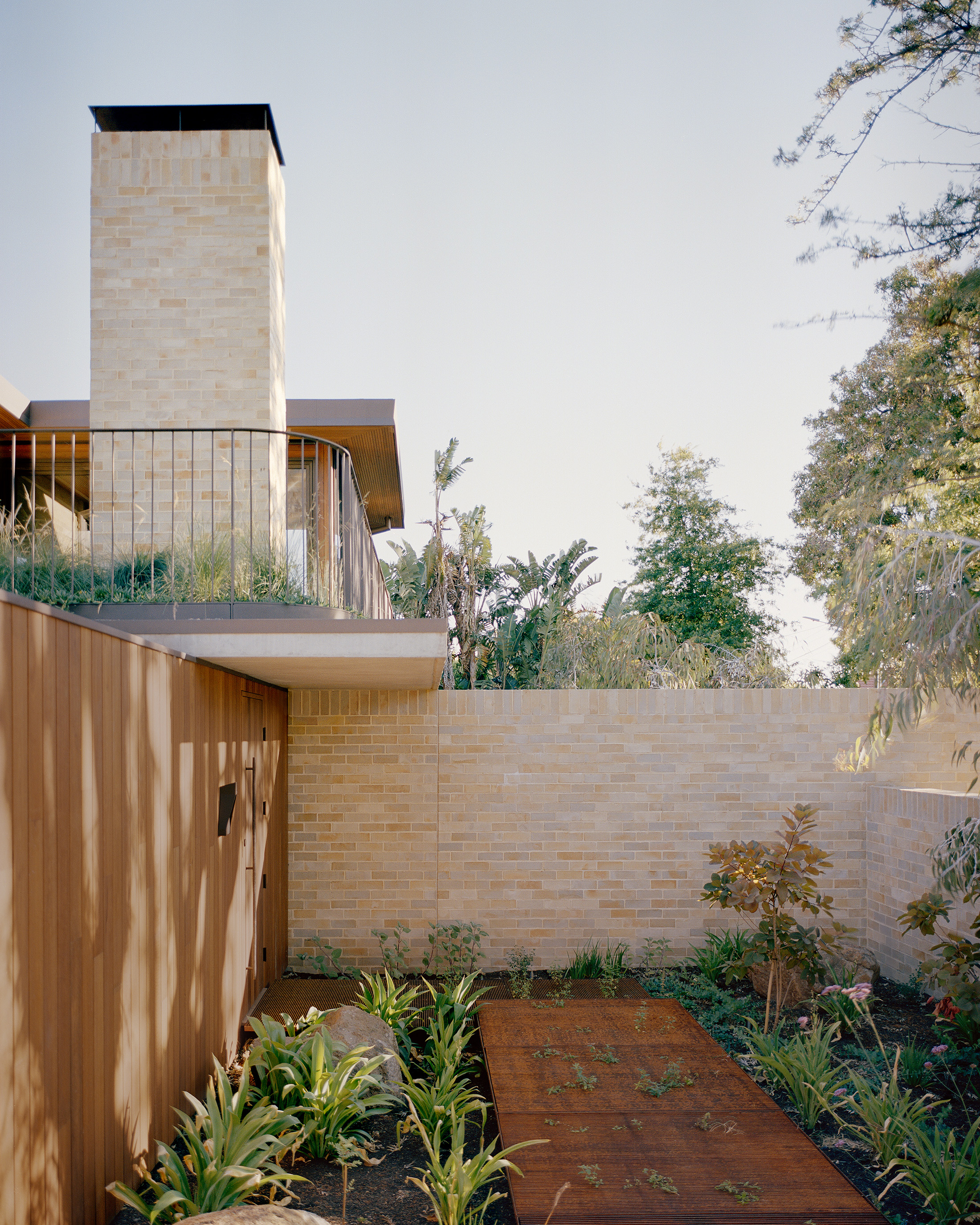 Modern landscape design featuring a sunken courtyard with green leafy plants and a rusted corten steel walkway. A light brick wall separates the space from a towering exterior brick chimney and an upper-level rooftop terrace with a steel balustrade.