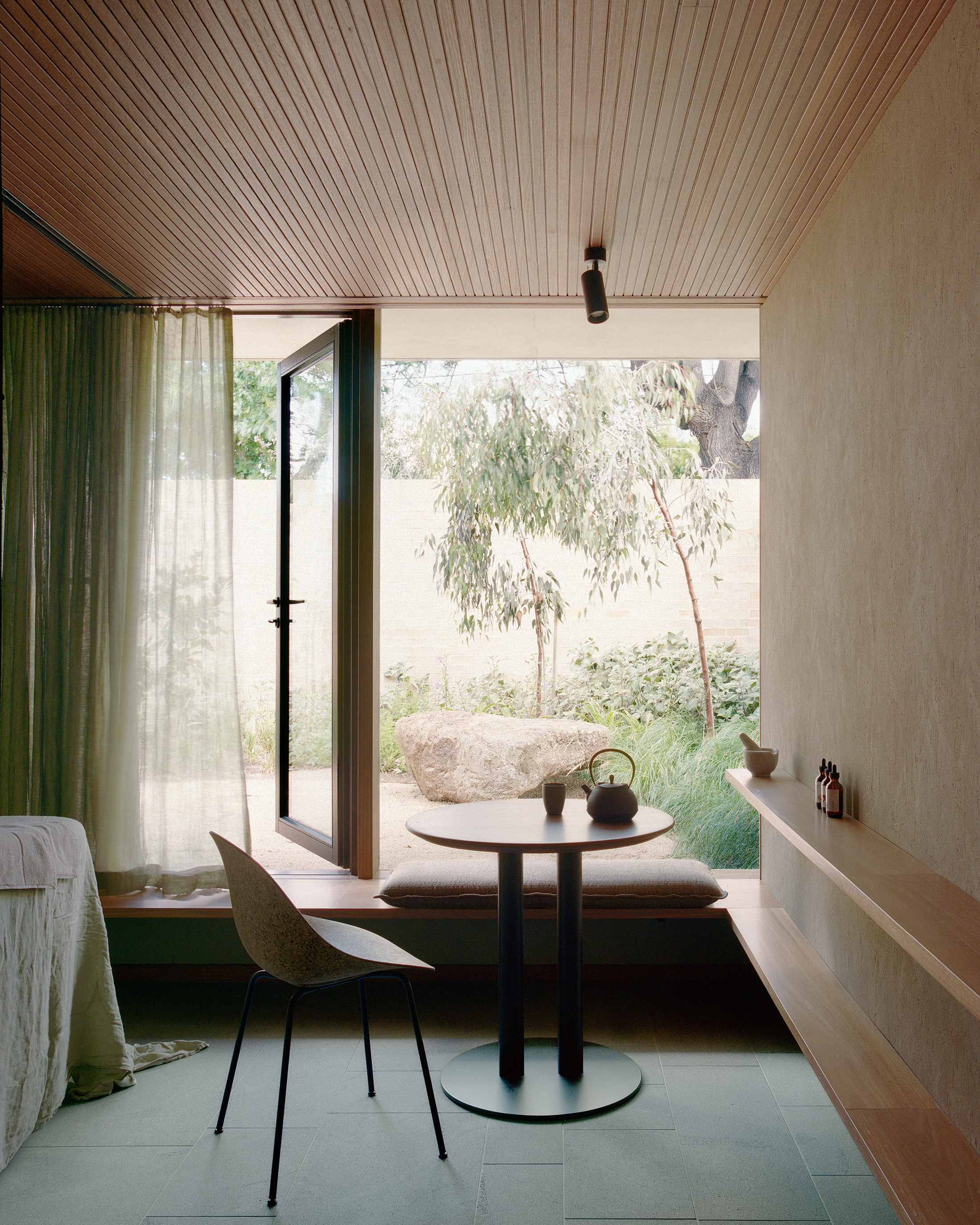 Minimalist wellness or treatment room interior featuring timber-lined ceilings, integrated timber shelving, and a small round tea table with a Japanese teapot. A glass door opens onto a serene native courtyard garden with a large natural boulder and eucaly
