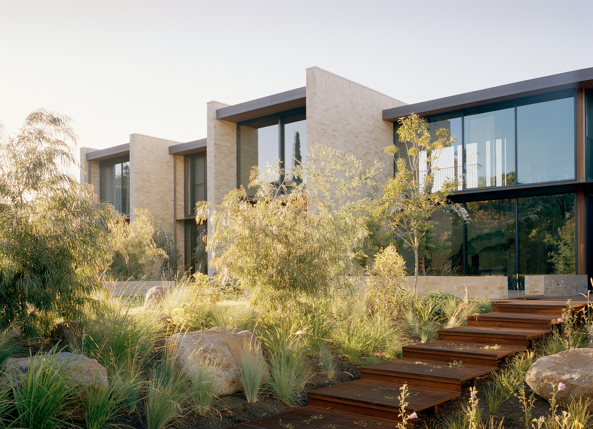 Contemporary multi-story brick townhouse design by Archier. The light-colored brick facade features floor-to-ceiling glass windows flanked by a sloping native garden path with weathered corten steel steps and eucalyptus trees.