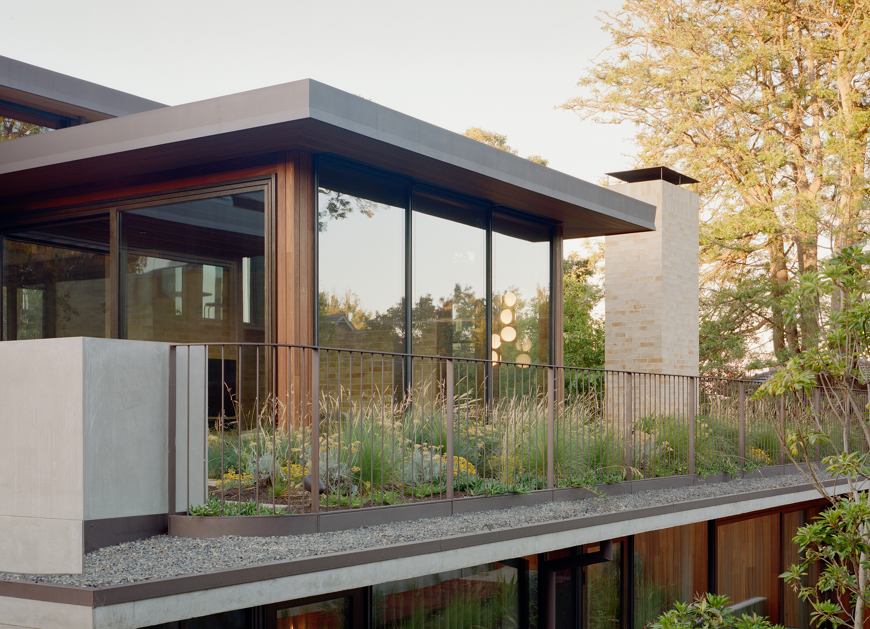 Modern rooftop terrace garden featuring wild grasses and native plantings bounded by a minimalist metal railing. The terrace connects to a timber and glass residential pavilion with an external brick chimney stack.