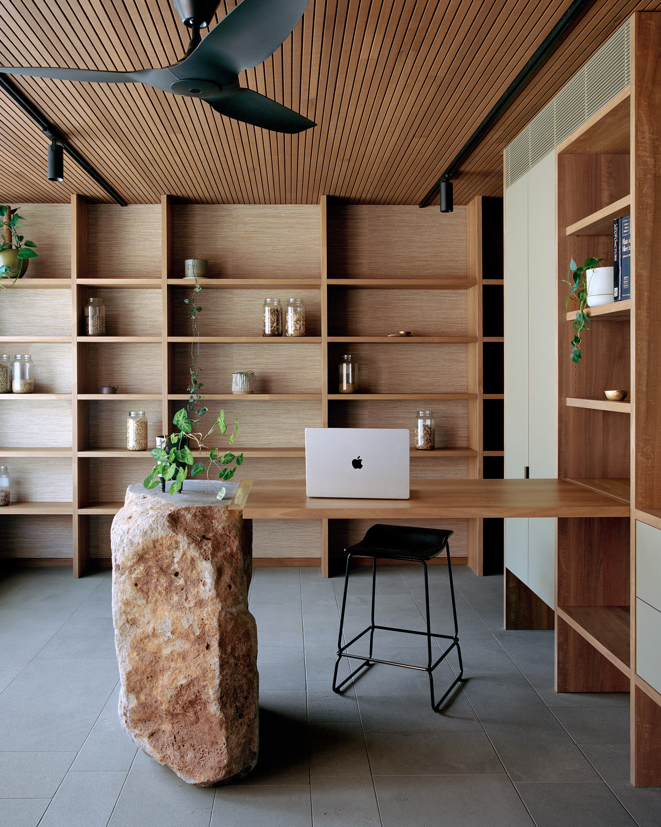 Contemporary office or consultation space designed by Archier. Features a custom timber desk supported by a massive natural stone boulder, a sleek black bar stool, a modern ceiling fan, and floor-to-ceiling wooden bookshelves filled with storage jars and p