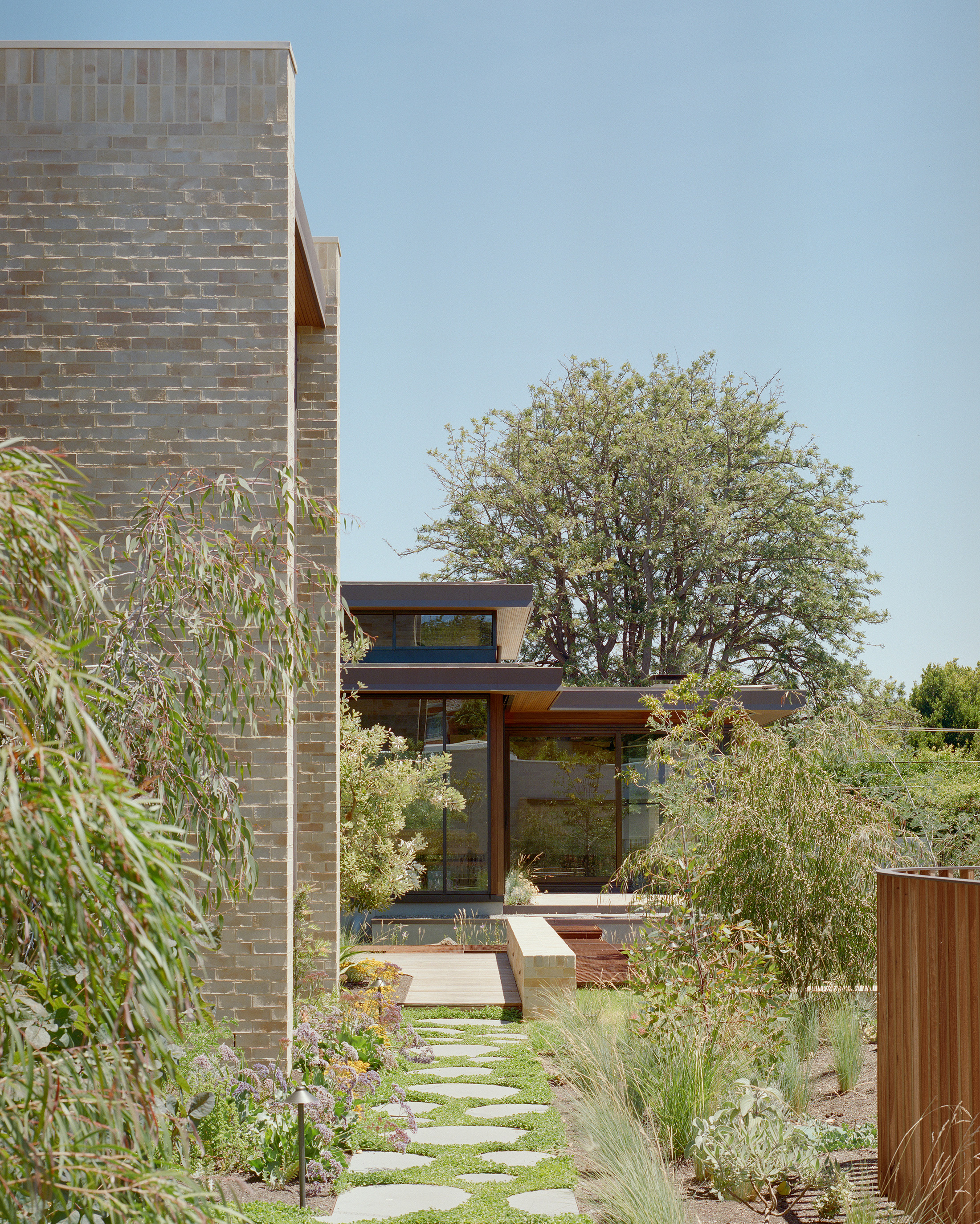 Residential landscape design featuring a stepping stone pathway set in green groundcover. The path leads past a light brick modern home wing toward a glass pavilion nestled under a large, mature deciduous tree.