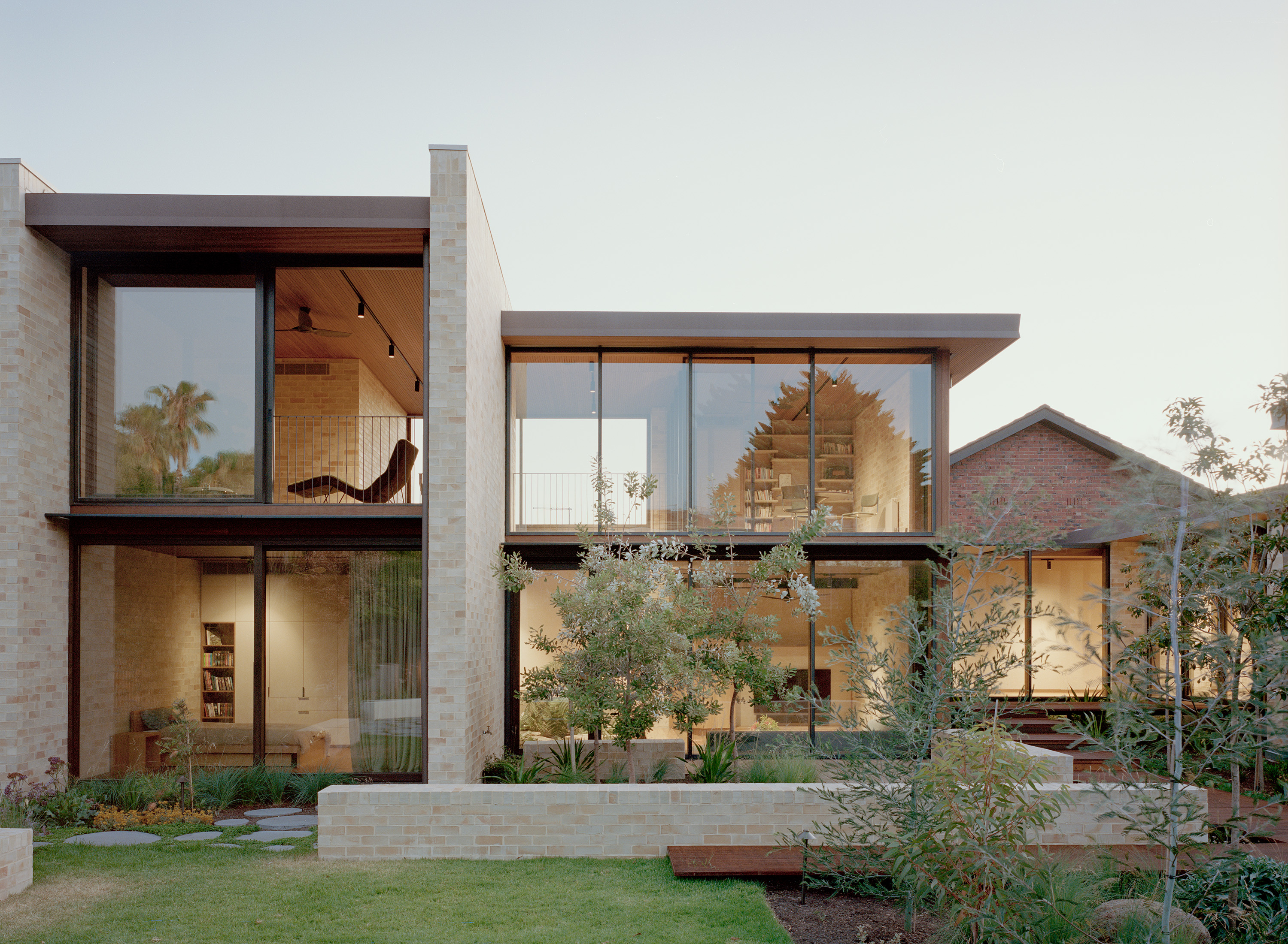Evening view of a luxury split-level modern home with extensive glazing, illuminating an internal home library and bedroom. The light brick structure integrates with a sunken courtyard garden and adjacent heritage red brick building.