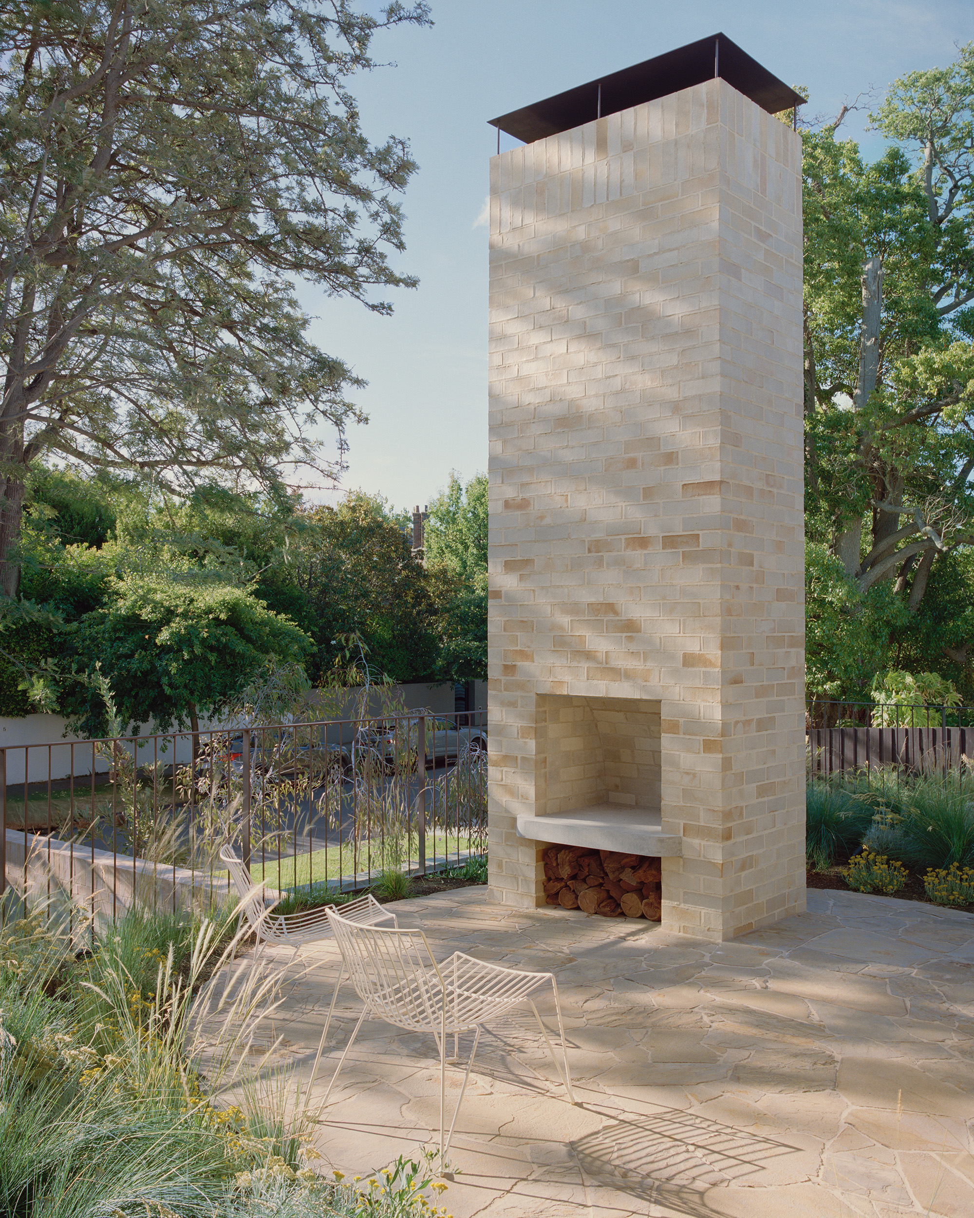 A minimalist outdoor brick fireplace and towering chimney on a flagstone patio. White wire-frame lounge chairs face the fireplace, integrated into a modern landscape design with native grasses and architectural steel fencing.