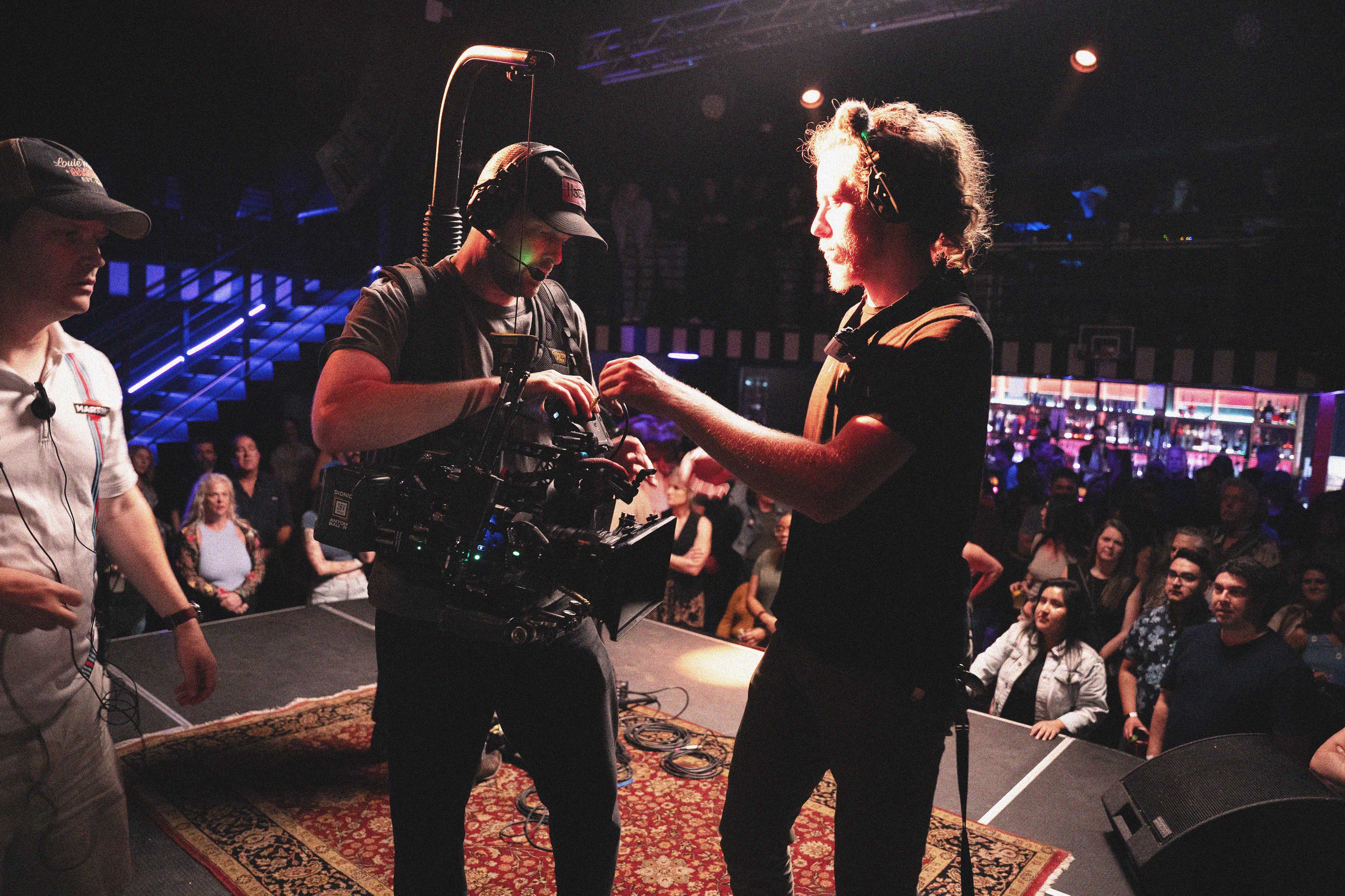 Two crew members with headsets preparing camera equipment on a stage with an audience in the background.