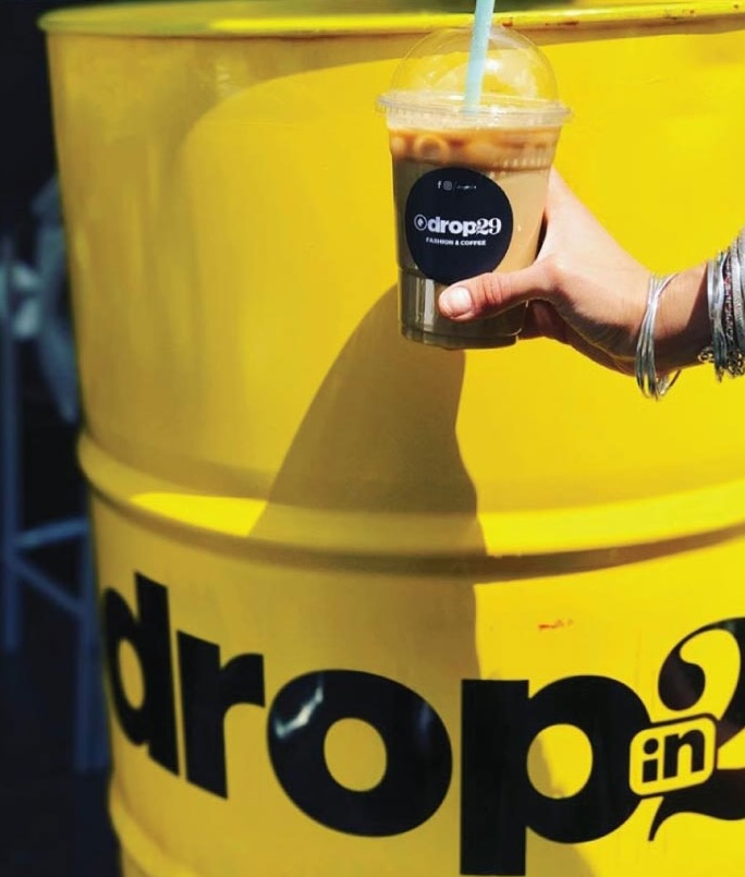 Close-up photo of an iced coffee held in front of Dropin29's yellow branding — placeholder, replace with Figma export