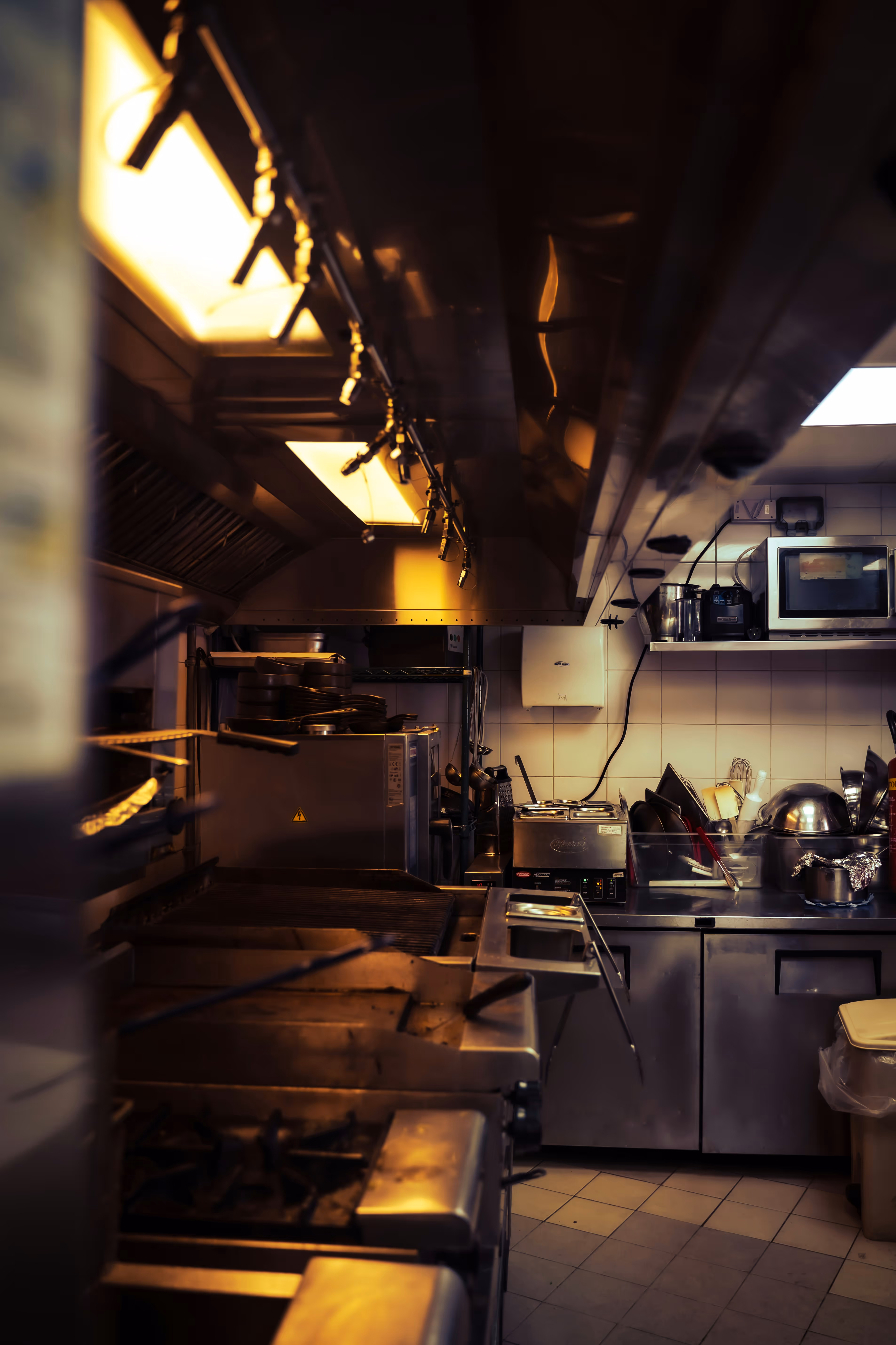 Dimly lit commercial kitchen with stainless steel appliances, cooking equipment, and utensils.