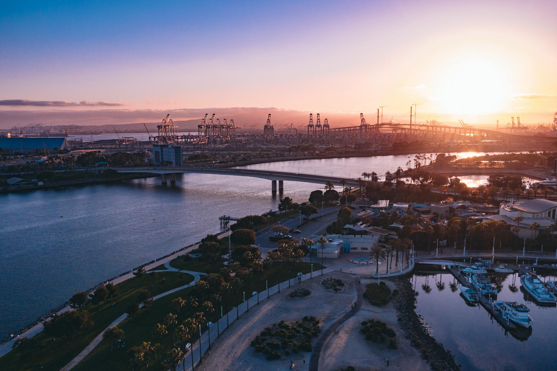 A serene sunset over a bustling harbor with bridges, cranes and tranquil waters.