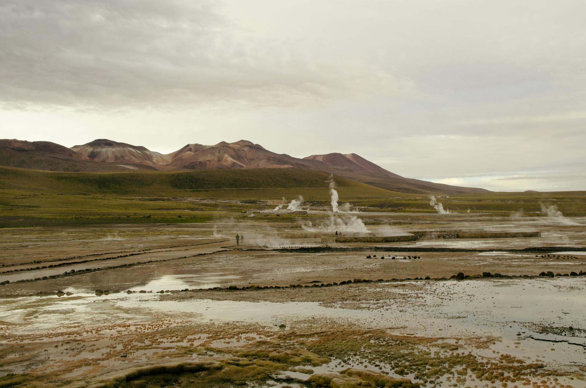 A dramatic scene of geysers emitting steam in a barren landscape with distant mountains.