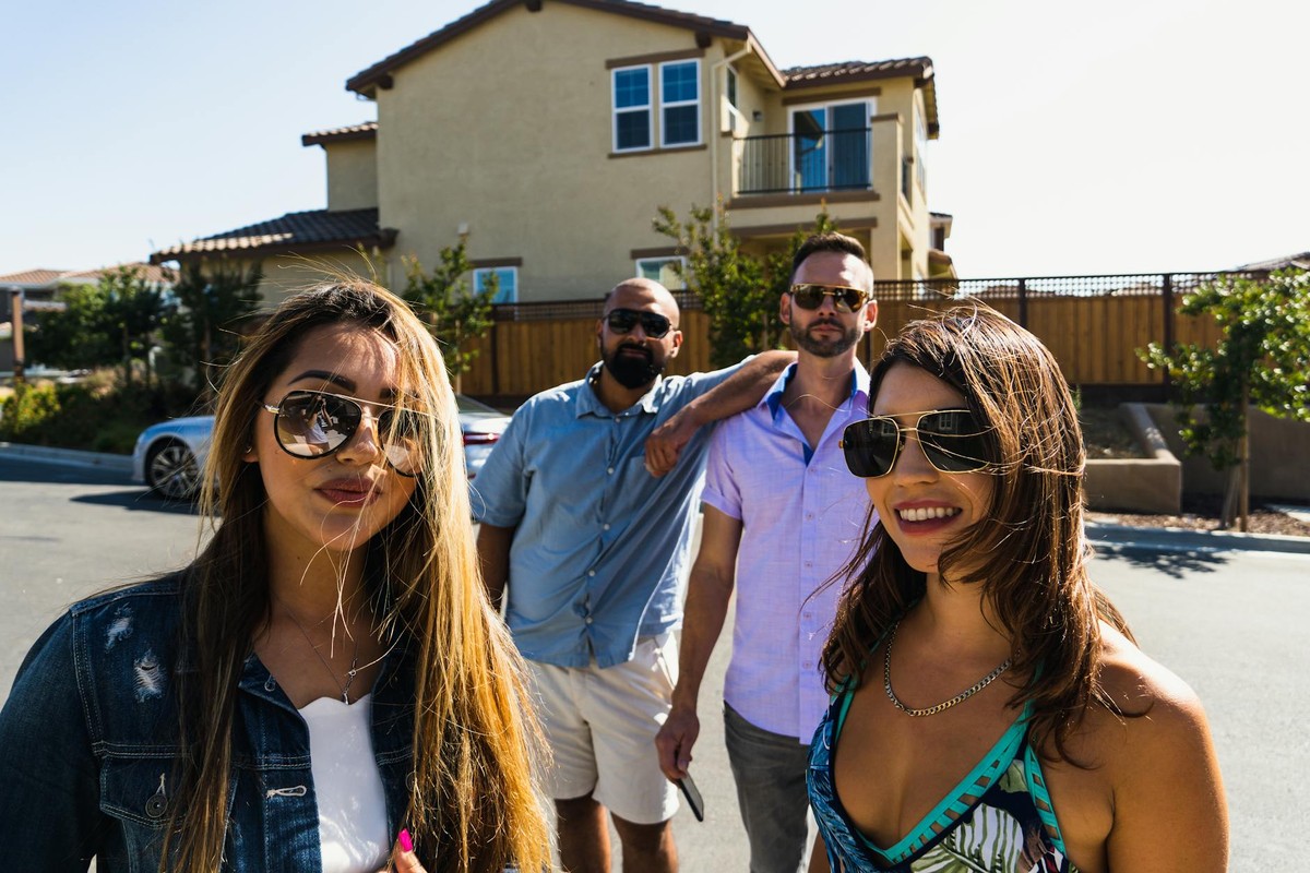 A group of adults smiling and posing outside a suburban house on a sunny day.