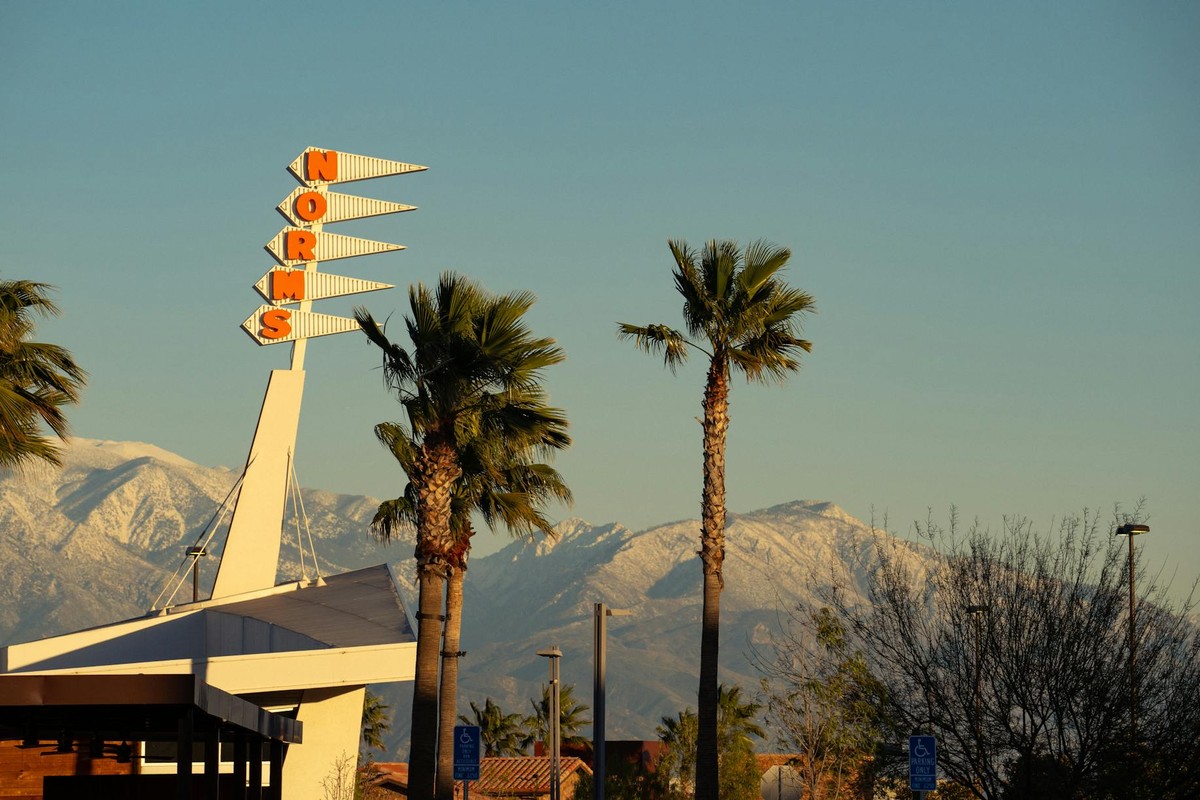 A retro Norms restaurant sign framed by palm trees and snow-capped mountains in Rialto, CA.