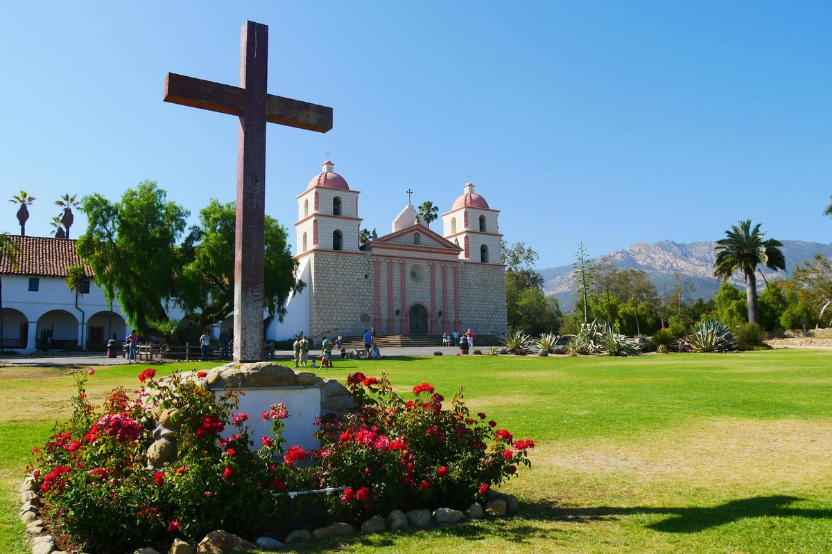 A sunny view of the Santa Barbara Mission with a cross and flowers in the foreground.
