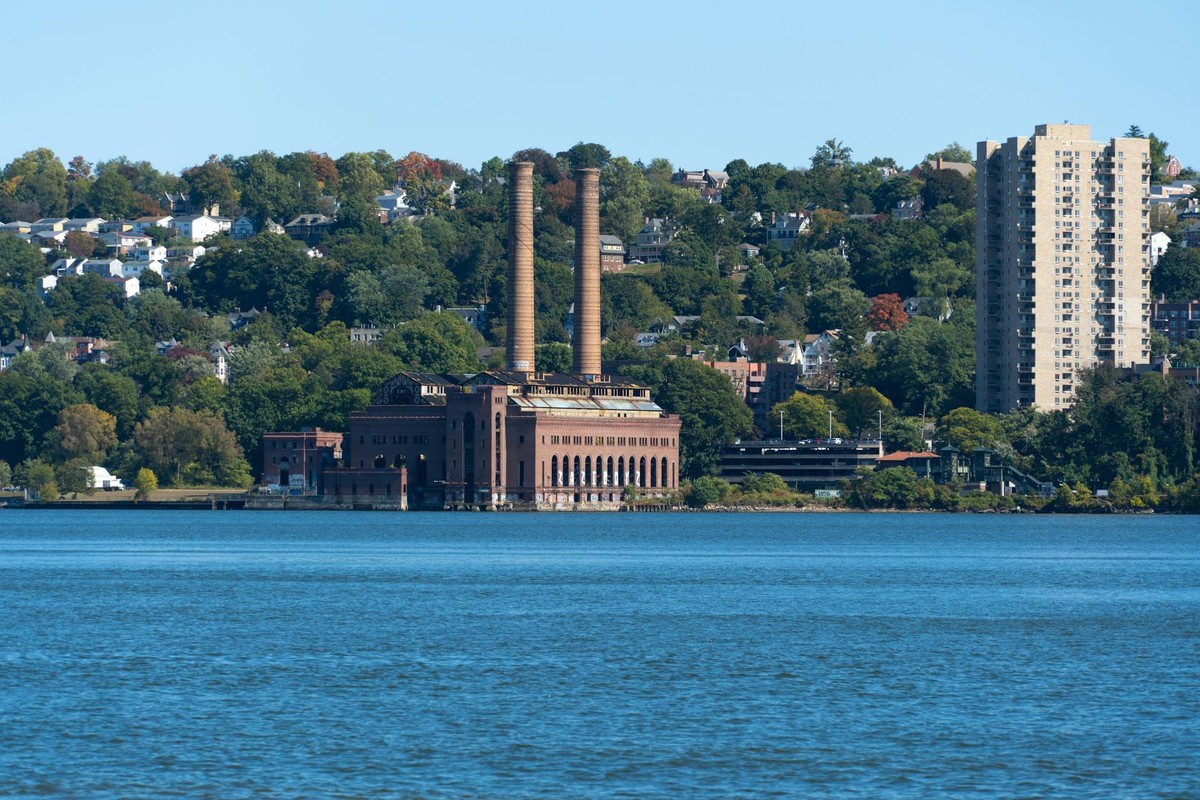 A historic industrial building with twin chimneys by a river with fall foliage and urban skyline.