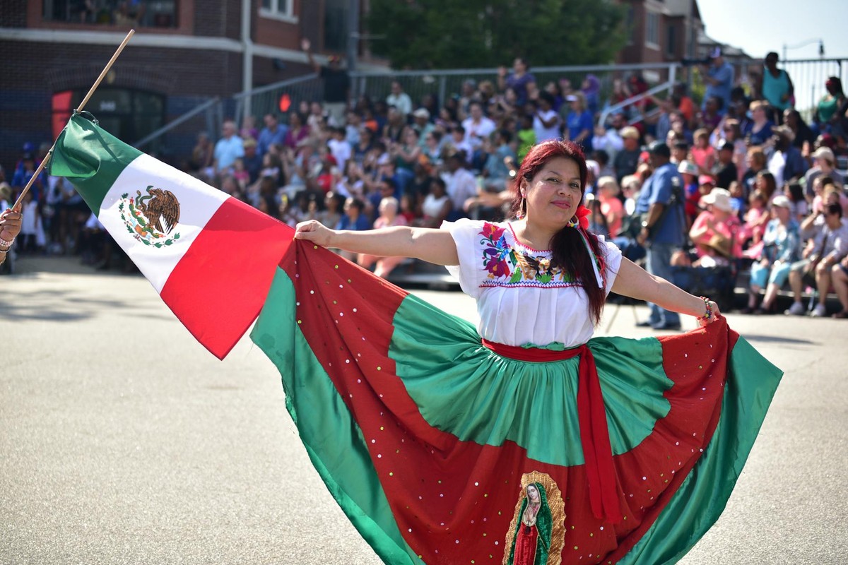 A woman in traditional Mexican attire dances with a Mexican flag during a street parade in Fayetteville.