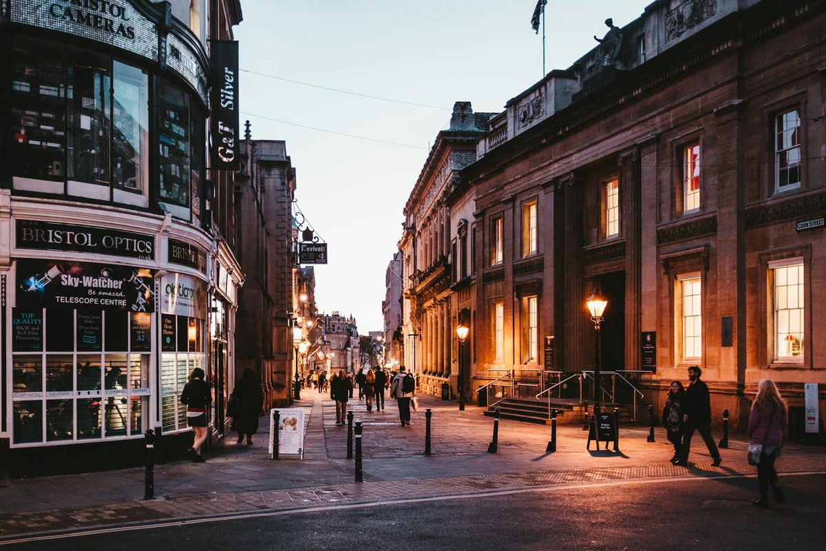 People walking along illuminated street in Bristol, England, during twilight with historic buildings.