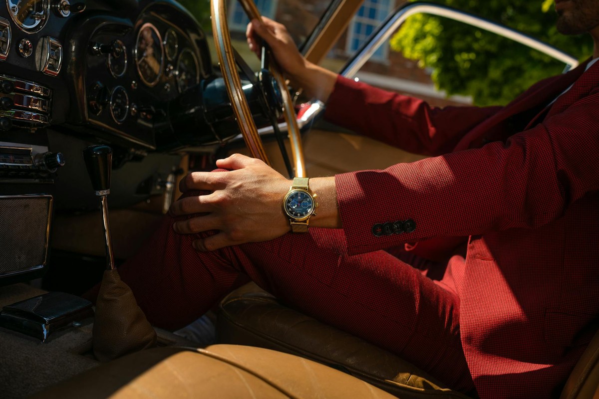 Man in red suit driving vintage car with gold watch and wooden steering wheel.