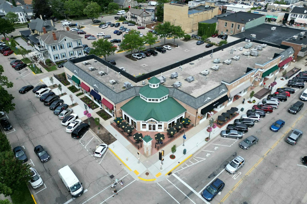 Bird's-eye view of a bustling suburban shopping center with parked cars and tree-lined streets.