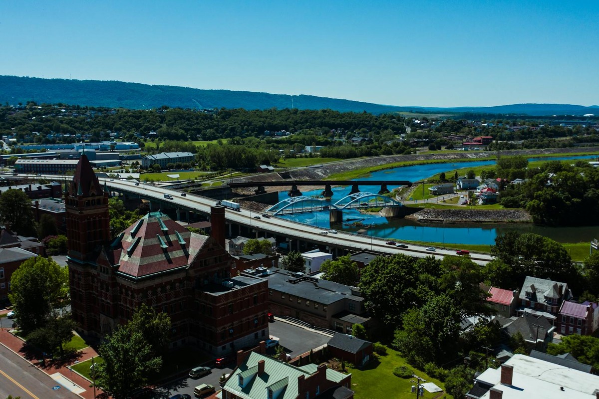 Drone view of contemporary bridge with vehicles above water channel near lawns and mounts under blue sky