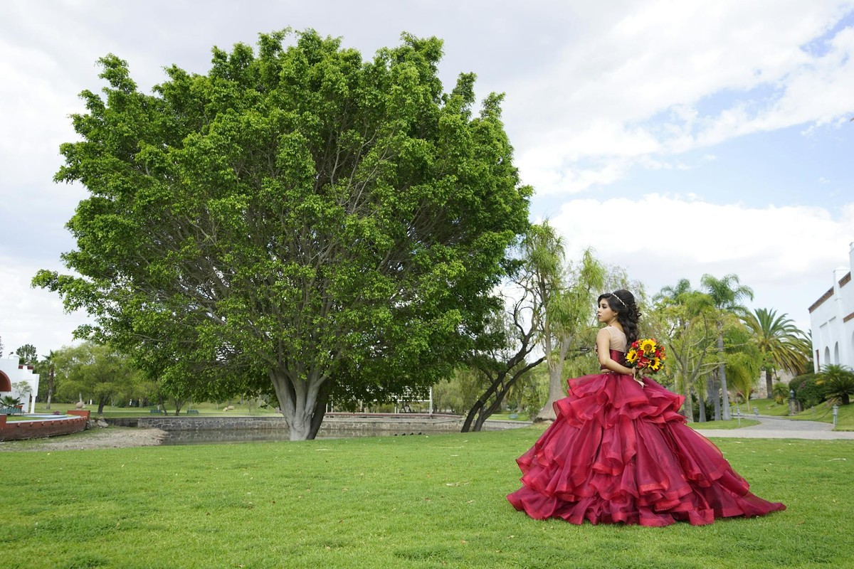 Young woman in red gown celebrating quinceañera outdoors in a lush garden setting.