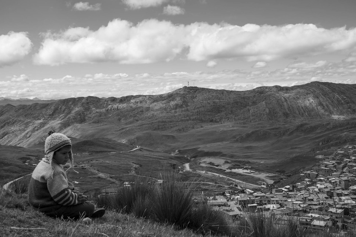 Child in traditional clothing sitting on a hill overlooking Cerro De Pasco's dramatic mountainous landscape.