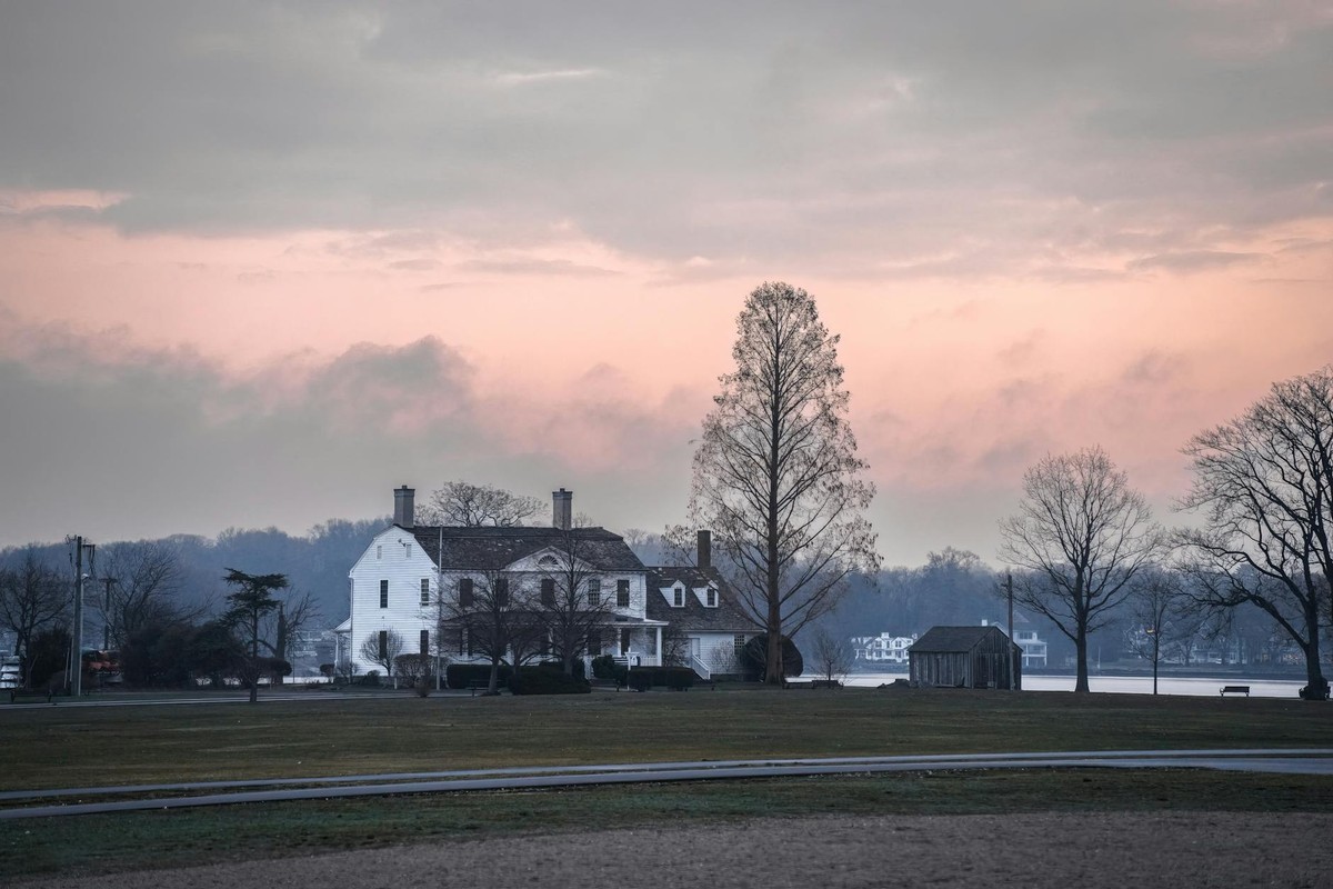 A picturesque historic house at sunset, surrounded by a tranquil landscape and bare trees.