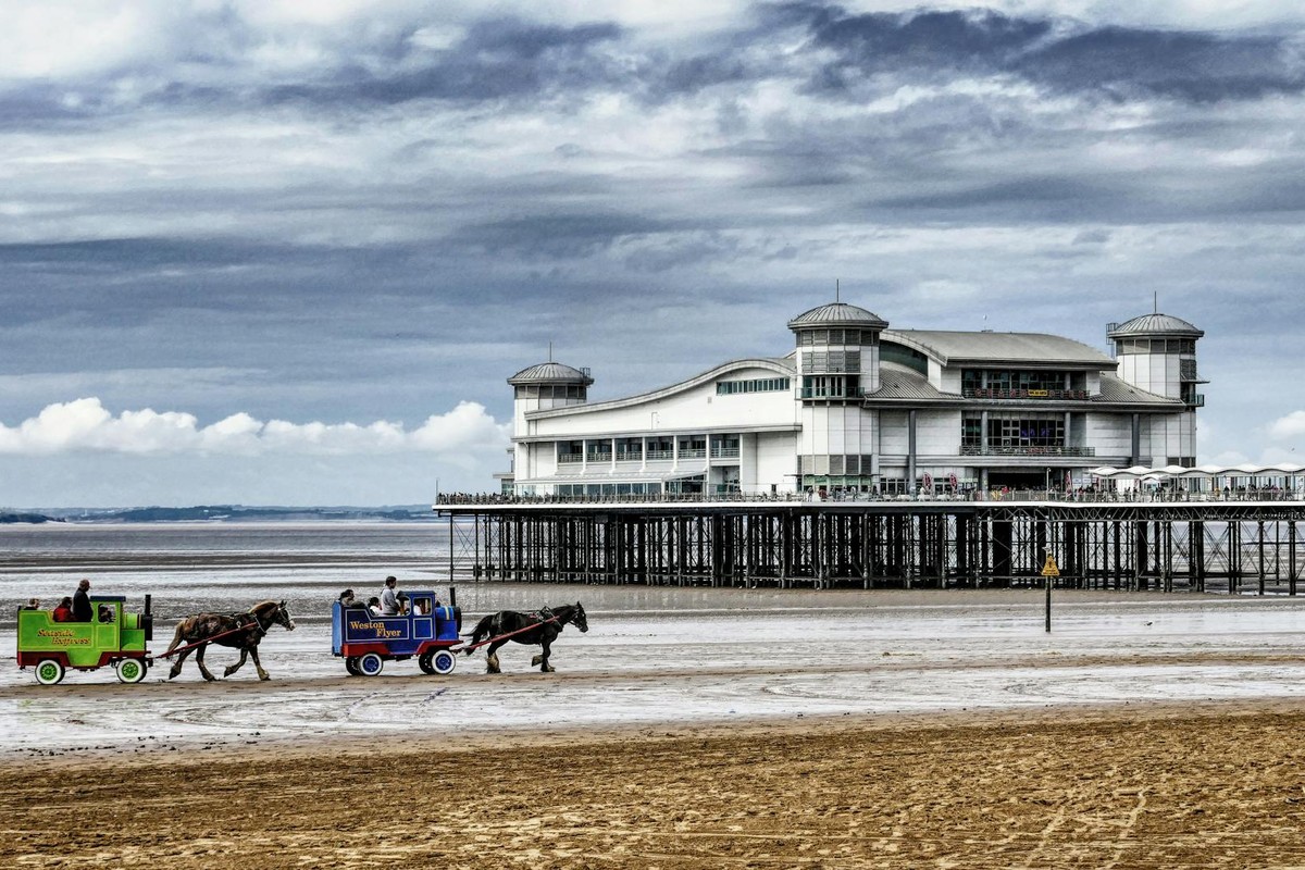Beautiful coastal scene with a pier, horse-drawn carriages, and moody clouds over the sea.