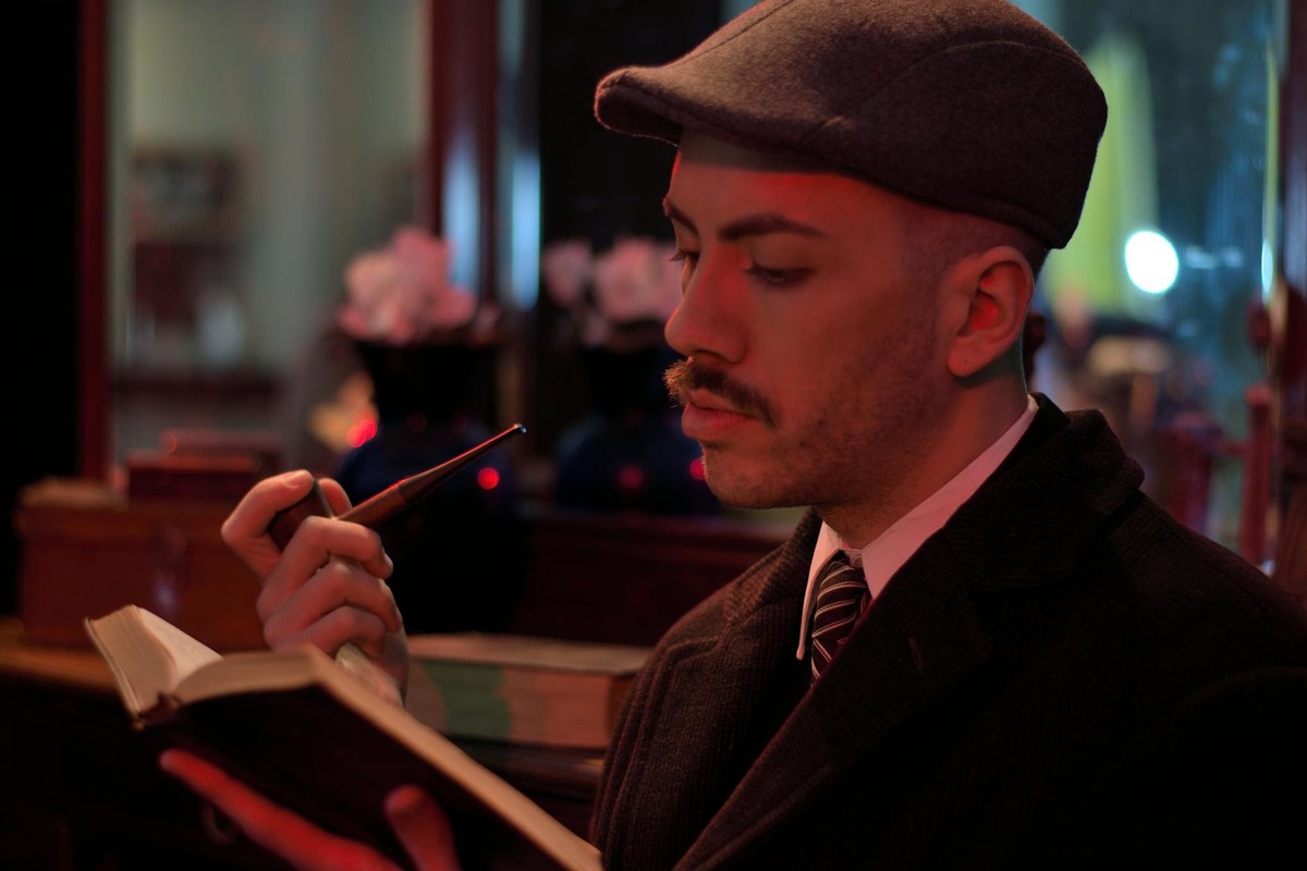 A man in vintage attire reading a book with a thoughtful expression in a cozy interior setting.